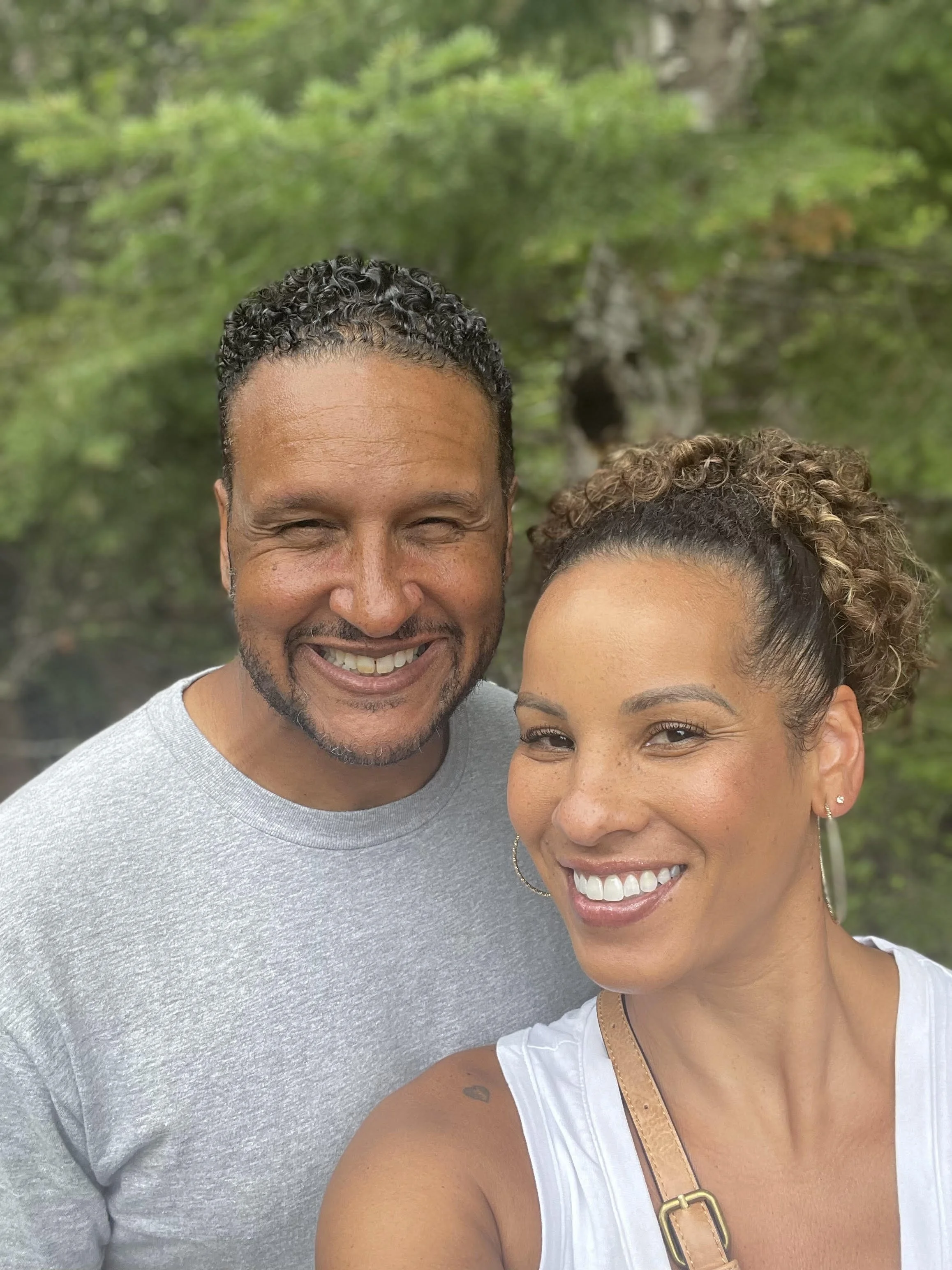 A smiling man and woman taking a selfie outdoors with a green, leafy background.