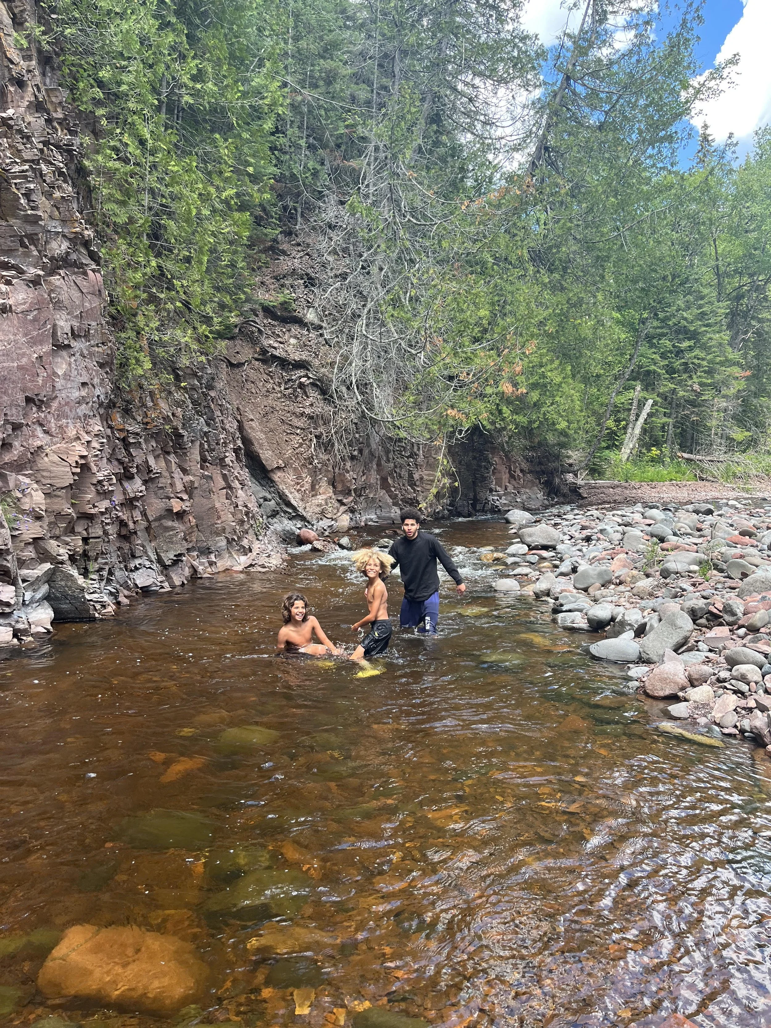 Three people playing in a shallow rocky river surrounded by trees and a cliff.