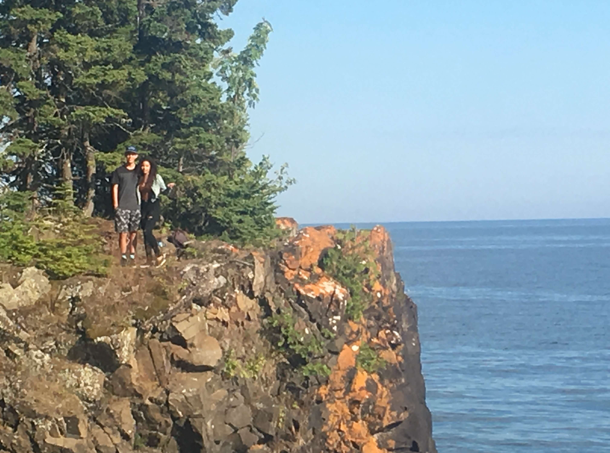 Two people standing on a rocky cliff near trees, overlooking a body of water on a sunny day.