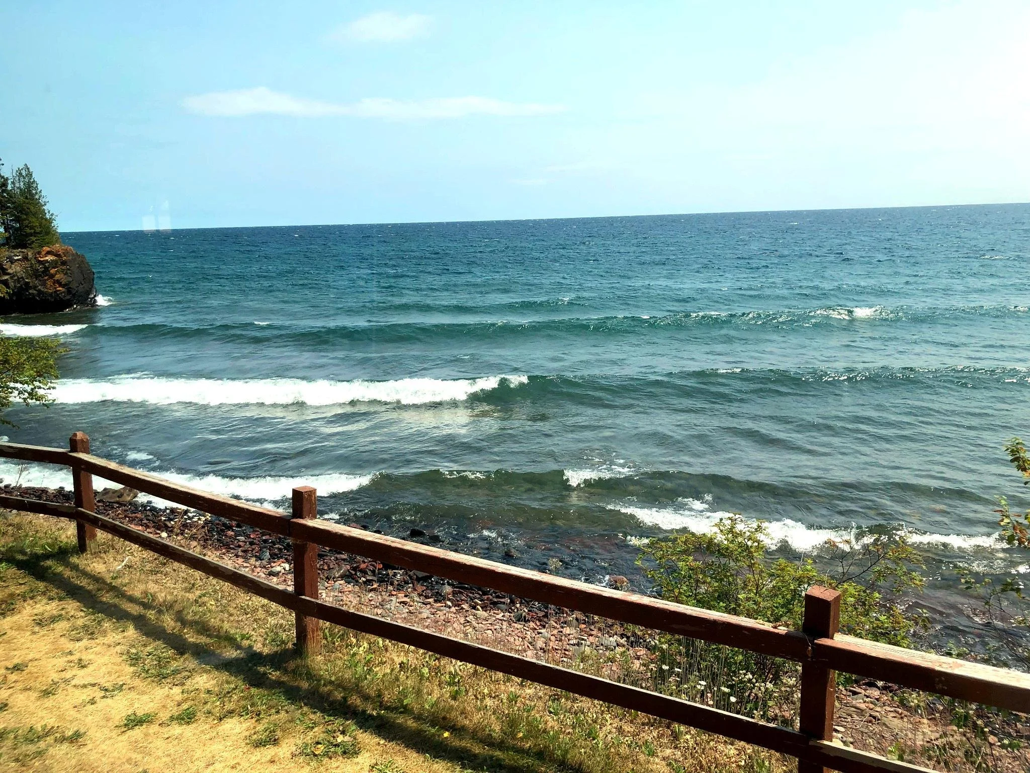 View of the ocean with waves hitting the shore, a wooden fence in the foreground, some greenery, and a rocky outcrop on the left side, under a partly cloudy sky.