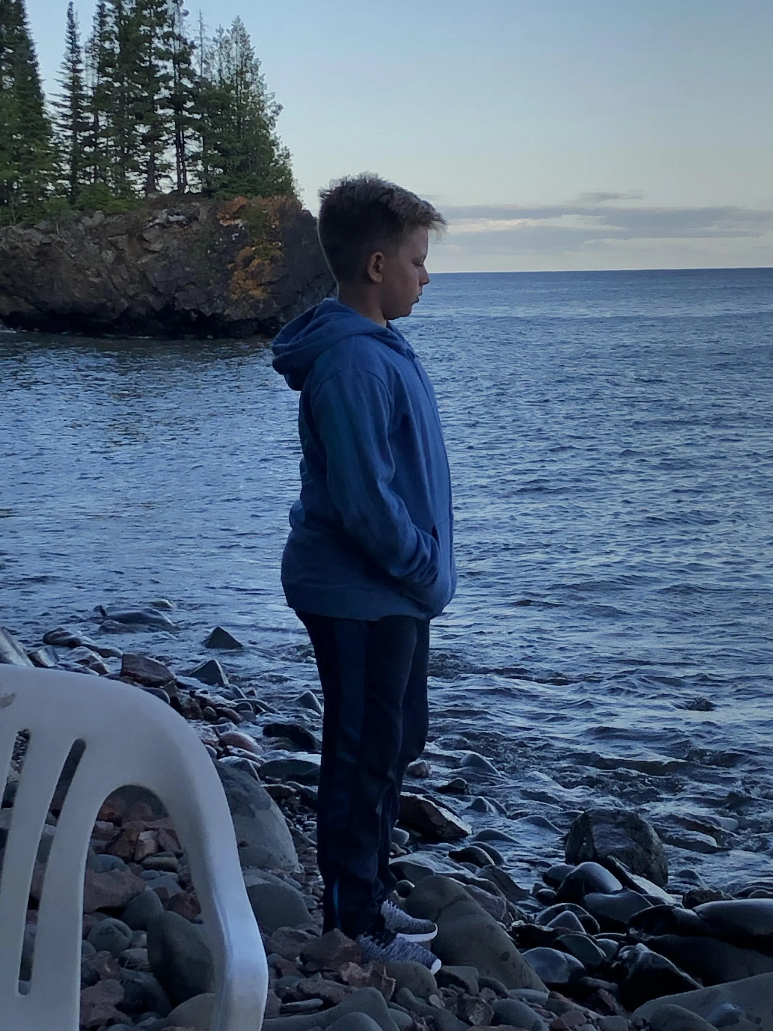 A boy standing on a rocky beach near the water, looking out at the ocean with trees on a rocky cliff in the background, wearing a blue hoodie and dark pants.