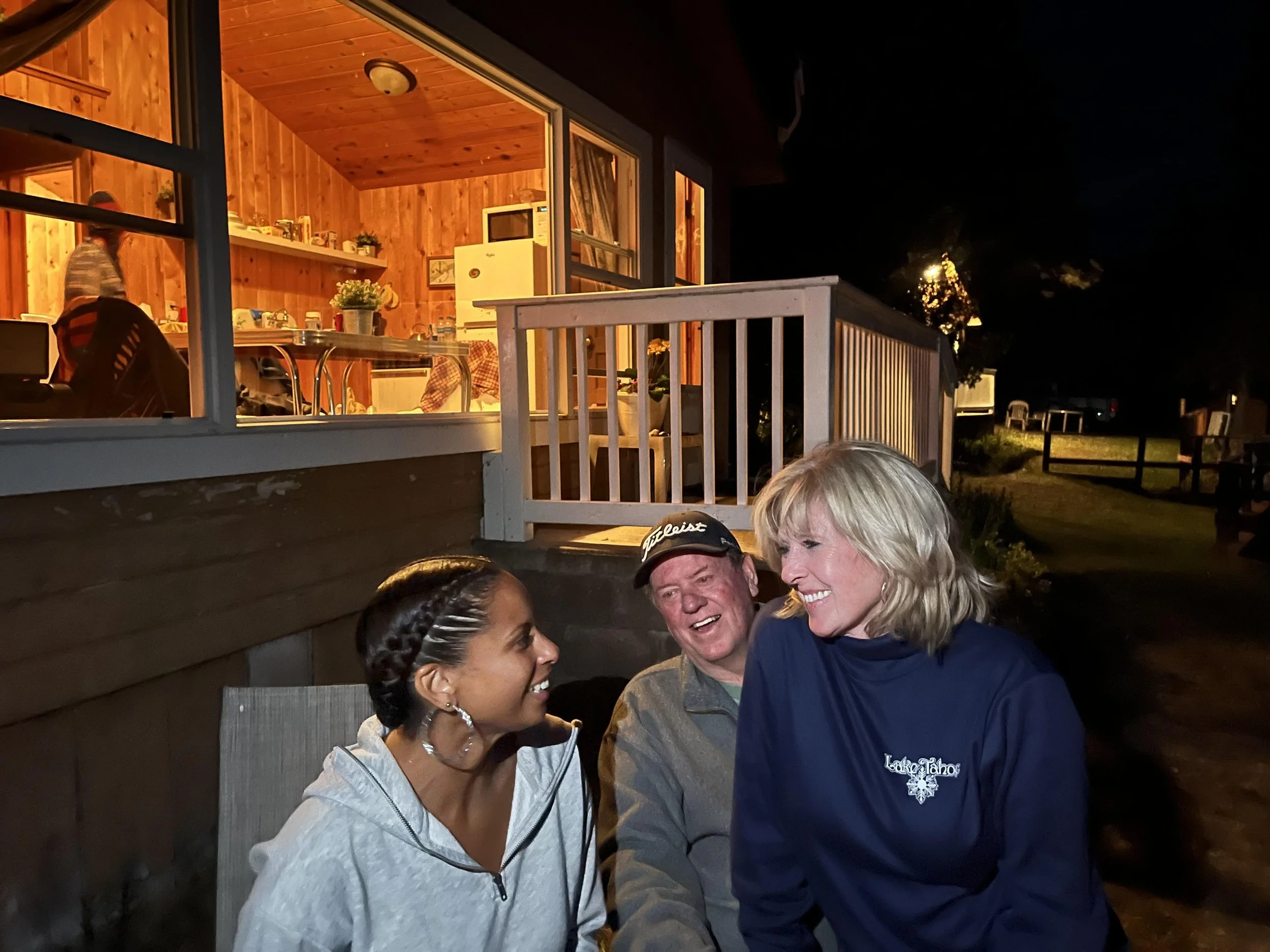 Three people sitting outside at night, smiling and talking, with a wooden house and lit interior visible behind them.