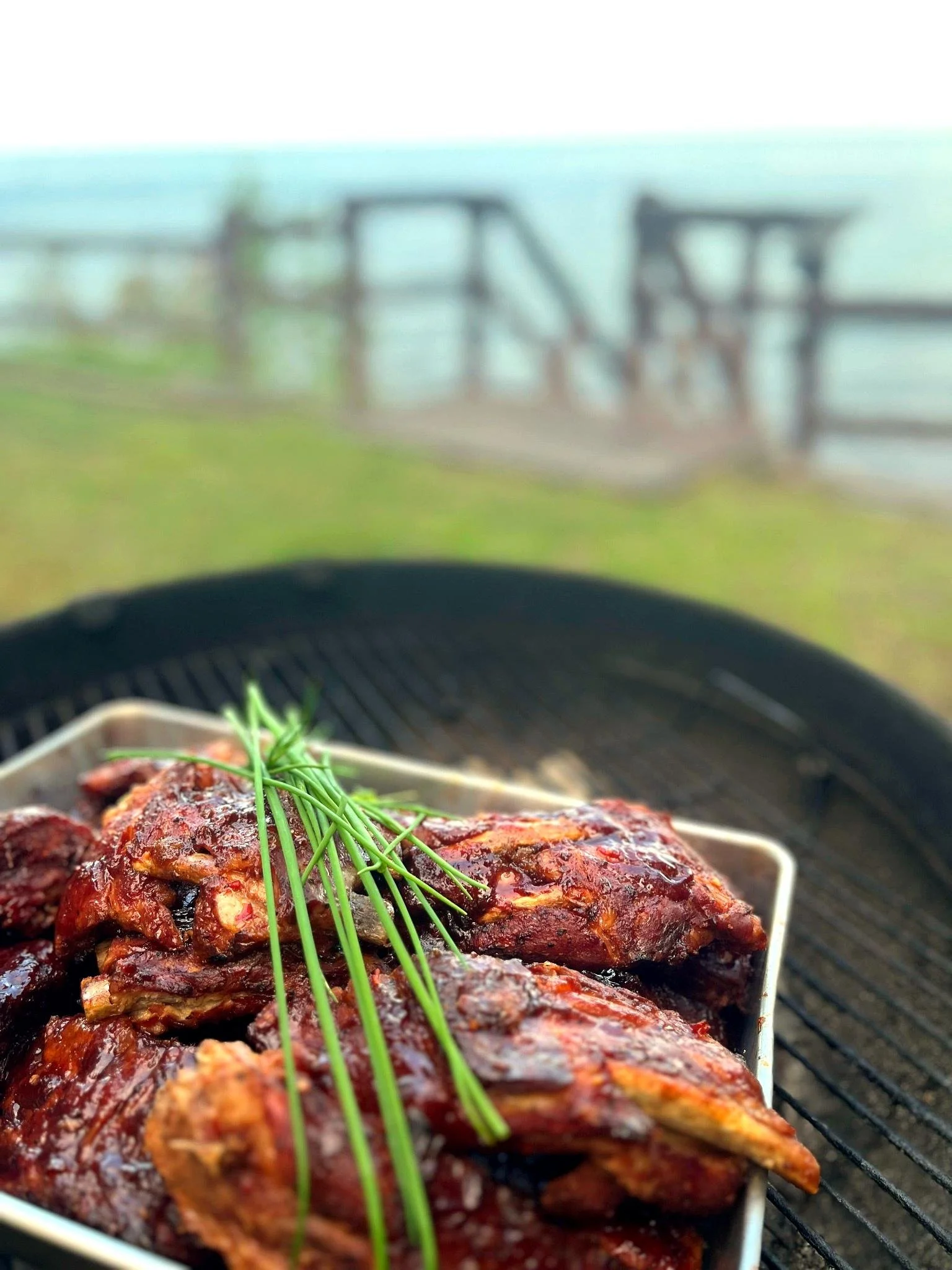 Barbecue ribs garnished with fresh chives on an outdoor grill with a foggy, outdoor setting in the background.