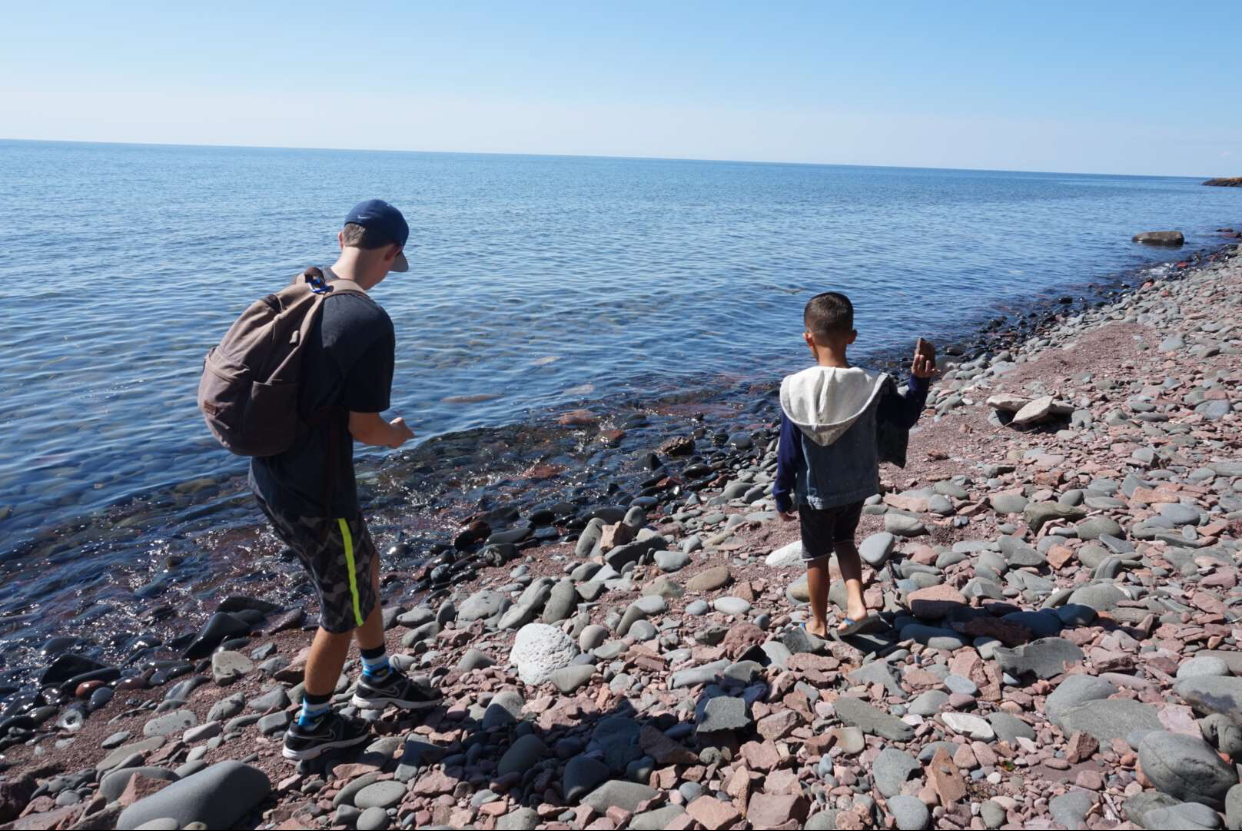 Two boys walking along a rocky shoreline by the water, with one holding a small item and the other carrying a backpack.