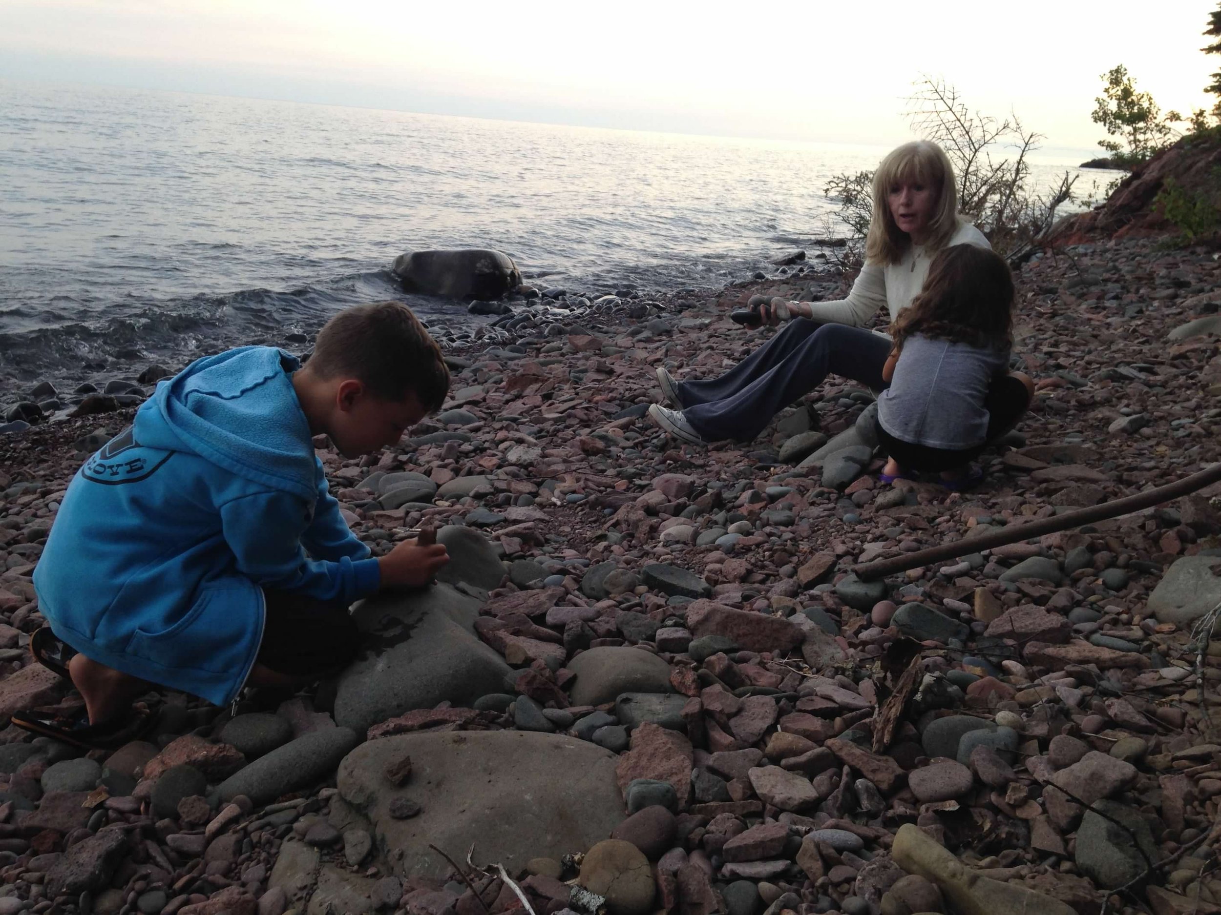 Three people sitting on a rocky lakeshore. One child in a blue hoodie is crouching and examining a rock. A woman with blonde hair, wearing a beige sweater and black pants, sits nearby. Another girl with brown hair, wearing a gray jacket, is sitting w