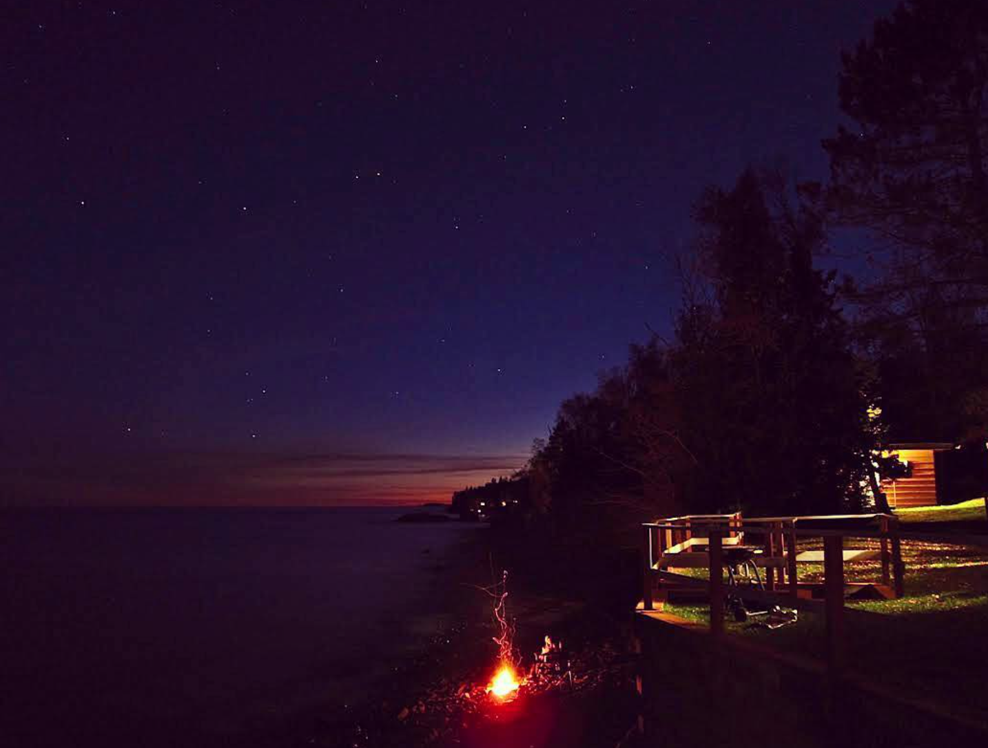 Nighttime scene of a lakeside with a bonfire, a wooden deck, and trees, under a starry sky.