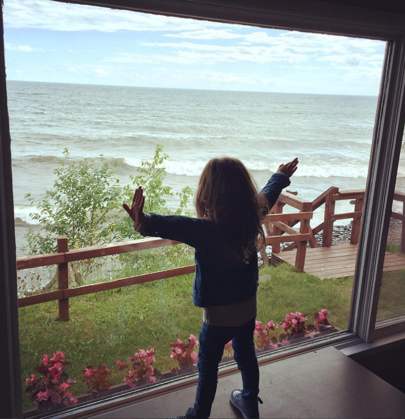Child standing inside and looking out at the ocean through a large window, with arms outstretched. Outside, there is a wooden deck, green grass, flowering plants, and waves on the water under a partly cloudy sky.