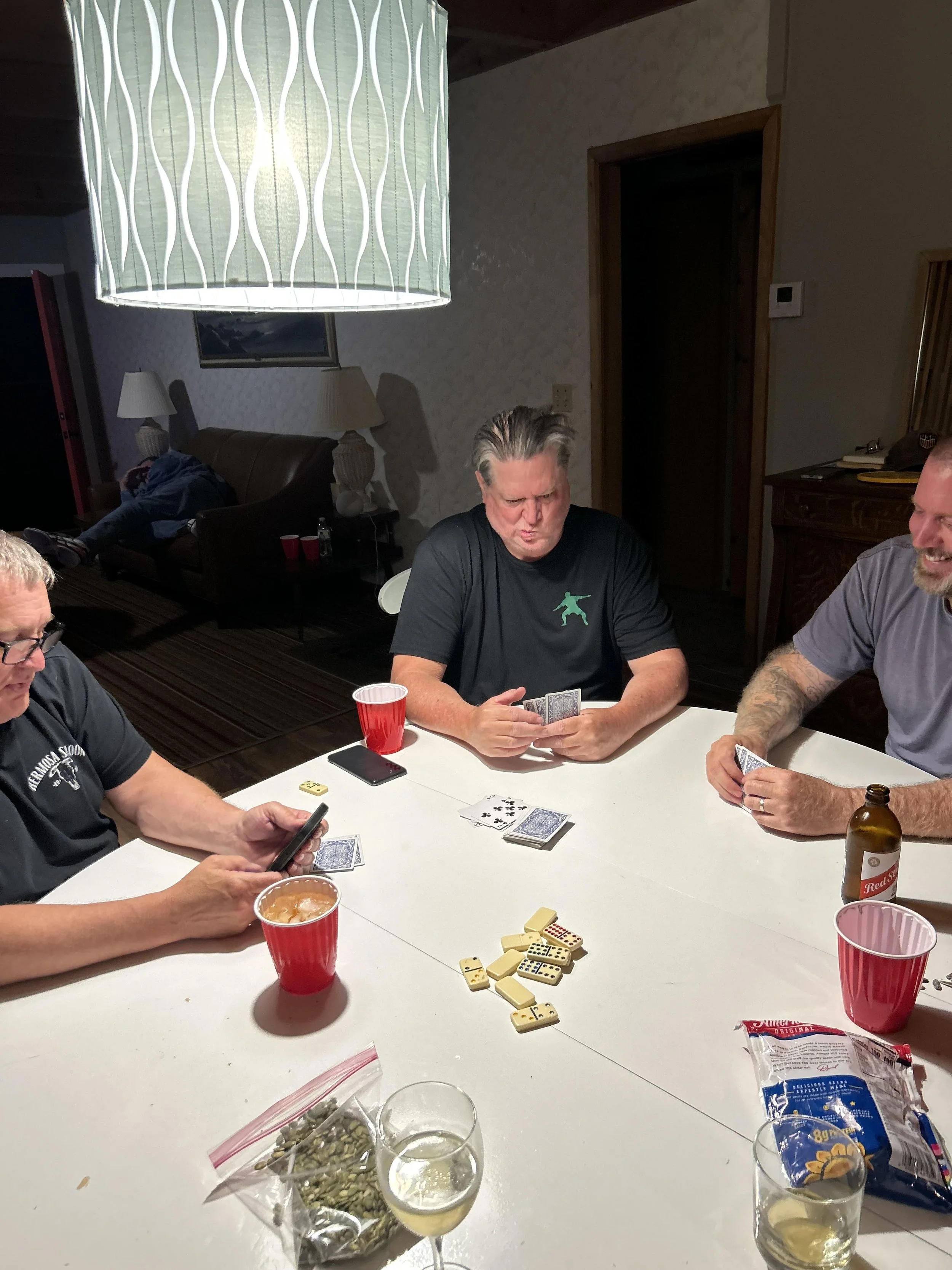 People playing a card game at a dining table, with snacks and drinks, in a casual indoor setting.
