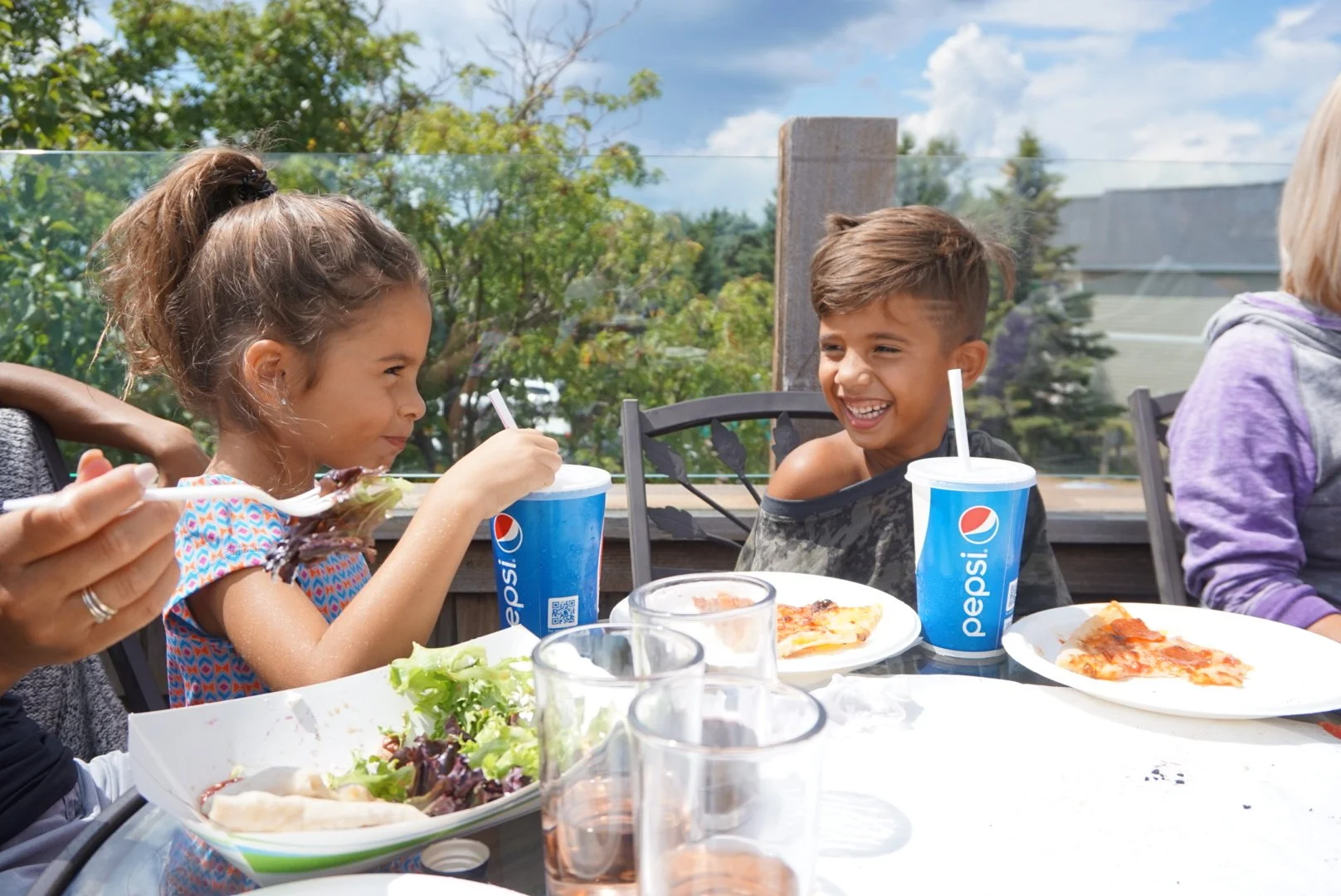 Children enjoying pizza and drinks at an outdoor table on a sunny day.