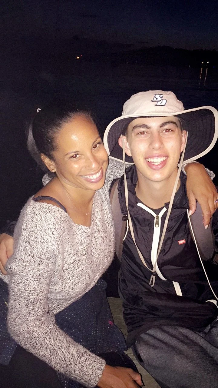 A woman and a young man smiling and posing for a photo at night near a body of water, with dark hills in the background.