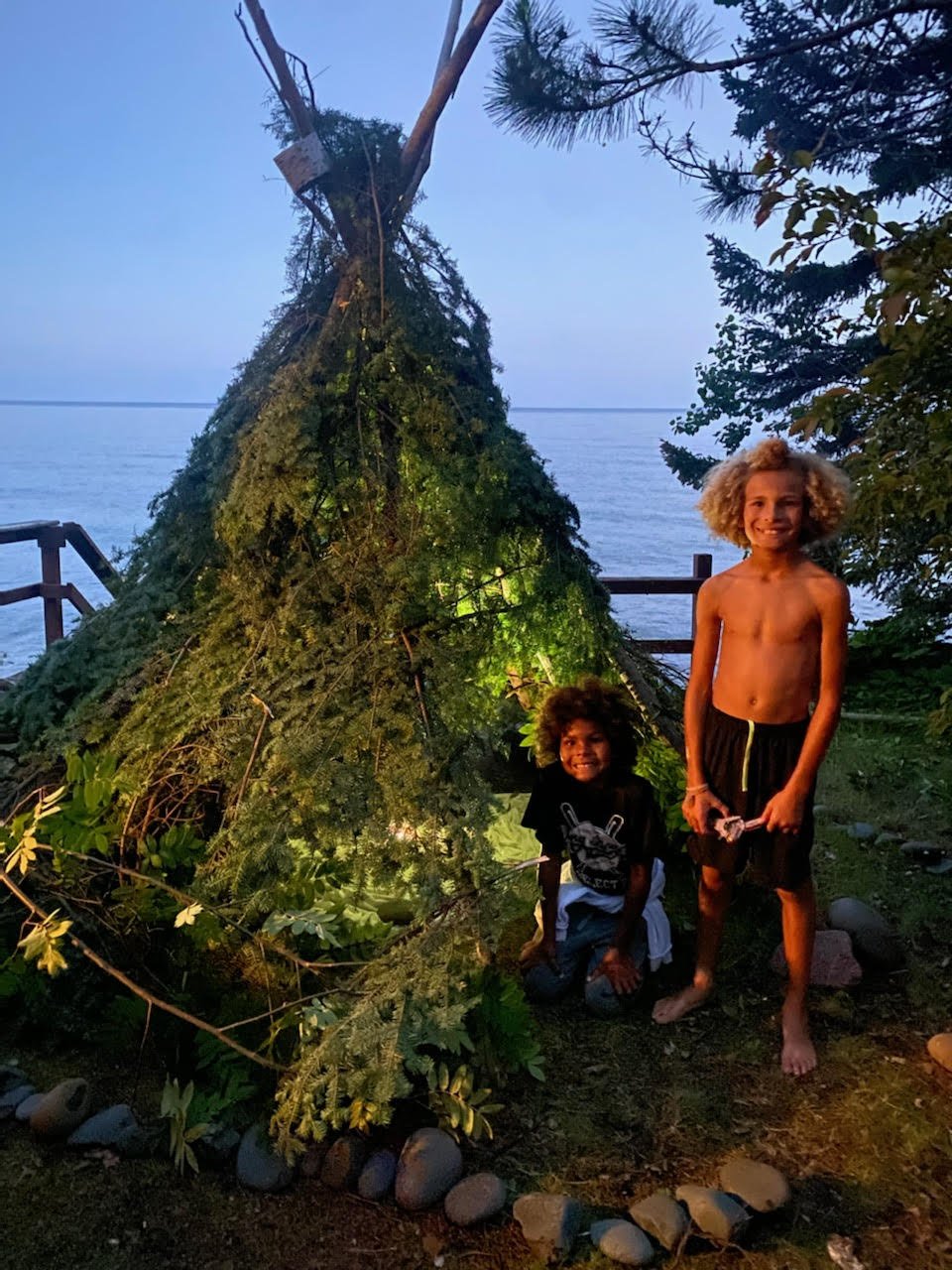 Two children with curly hair posing next to a small makeshift teepee made of branches and greenery near a body of water at dusk.