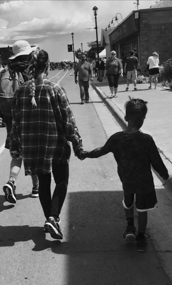 A woman and a young boy holding hands and walking down a street, with other people walking along the sidewalk and street shops nearby.