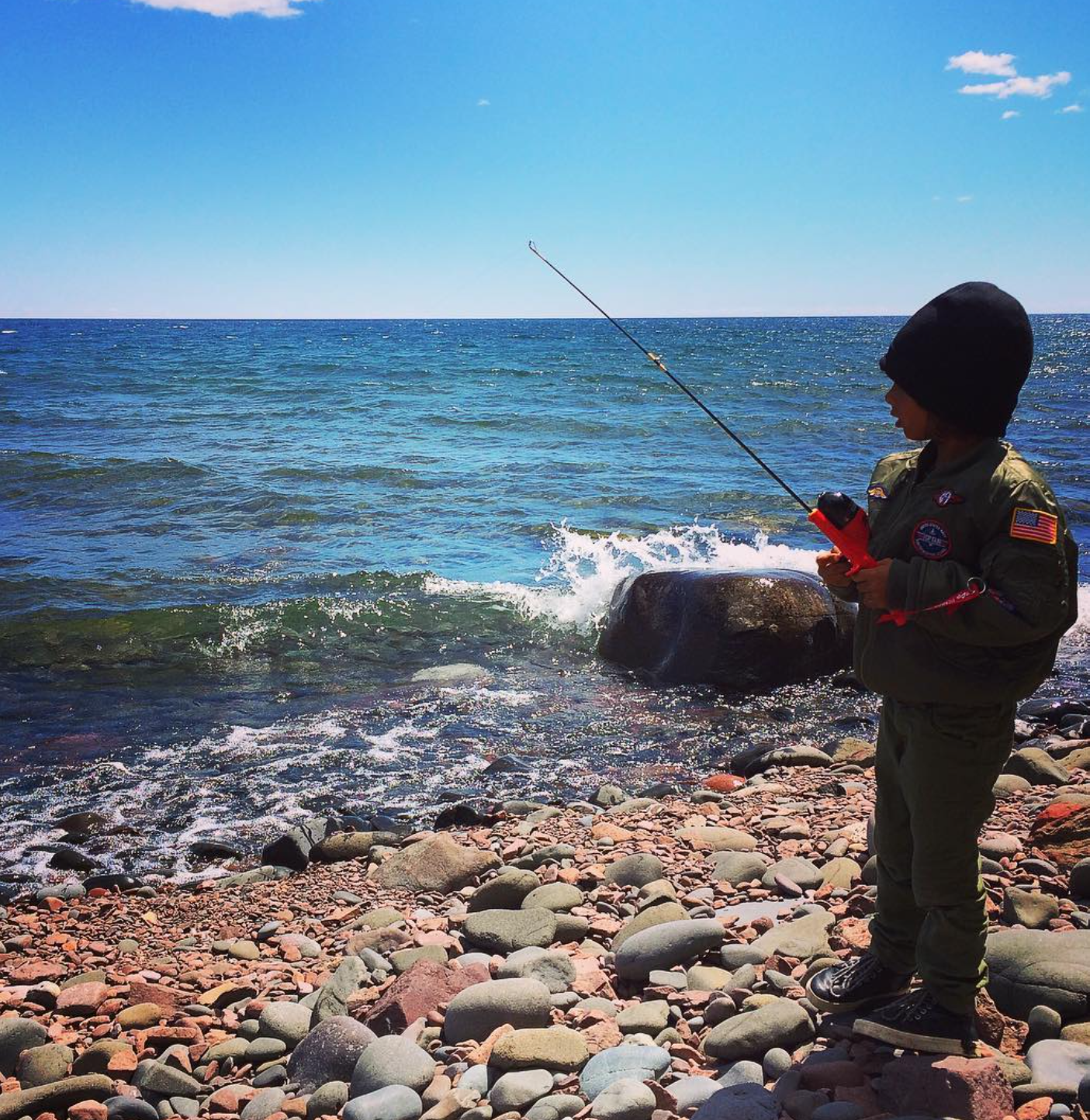 A person in a military or uniform jacket, wearing a beanie, standing on a rocky beach and fishing in the ocean with a rod during daytime.