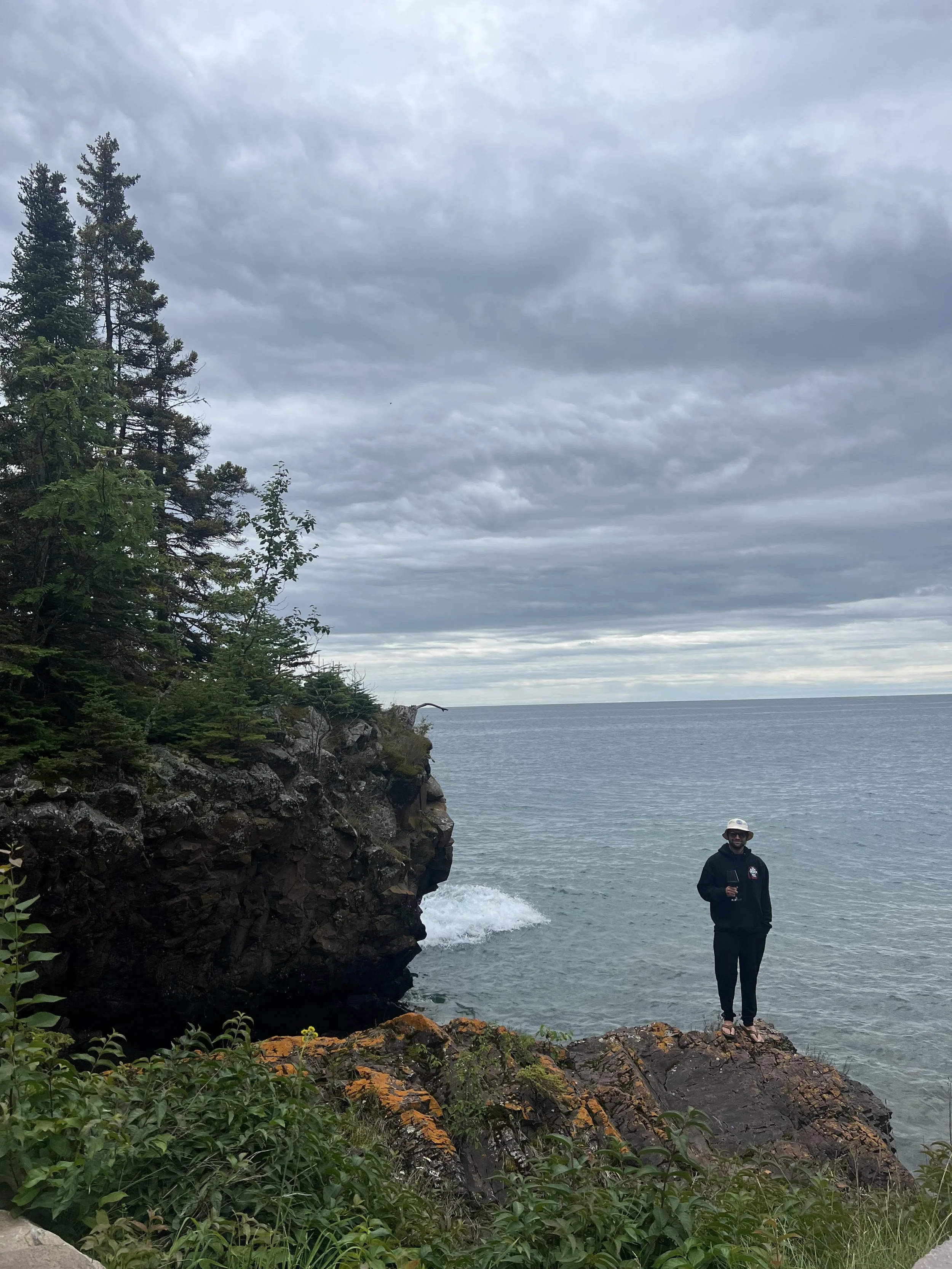 A person wearing a hat and dark clothing stands on rocks near the water at a shoreline, with trees on a rocky cliff to the left and a cloudy sky overhead.