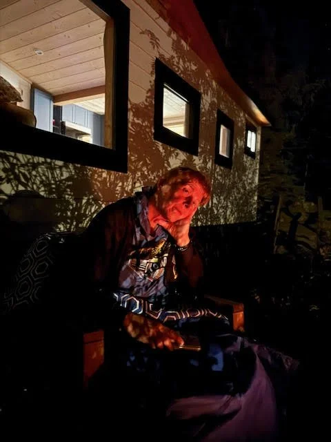 An elderly woman sitting outdoors at night, illuminated by a nearby light, with a house wall and windows in the background.