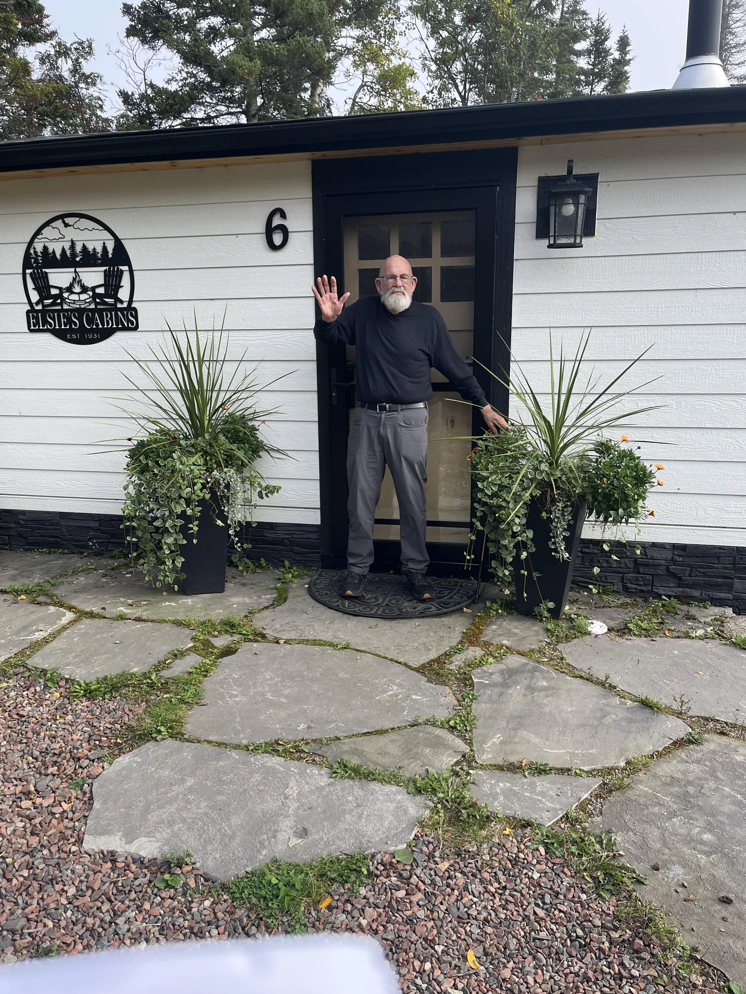A man with a beard, wearing glasses, a black long-sleeve shirt, and gray pants, is standing at the door of a cabin, waving with one hand. The cabin has a sign that reads 'Elsie's Cabins' with a mountain and forest logo. There are two large potted pla