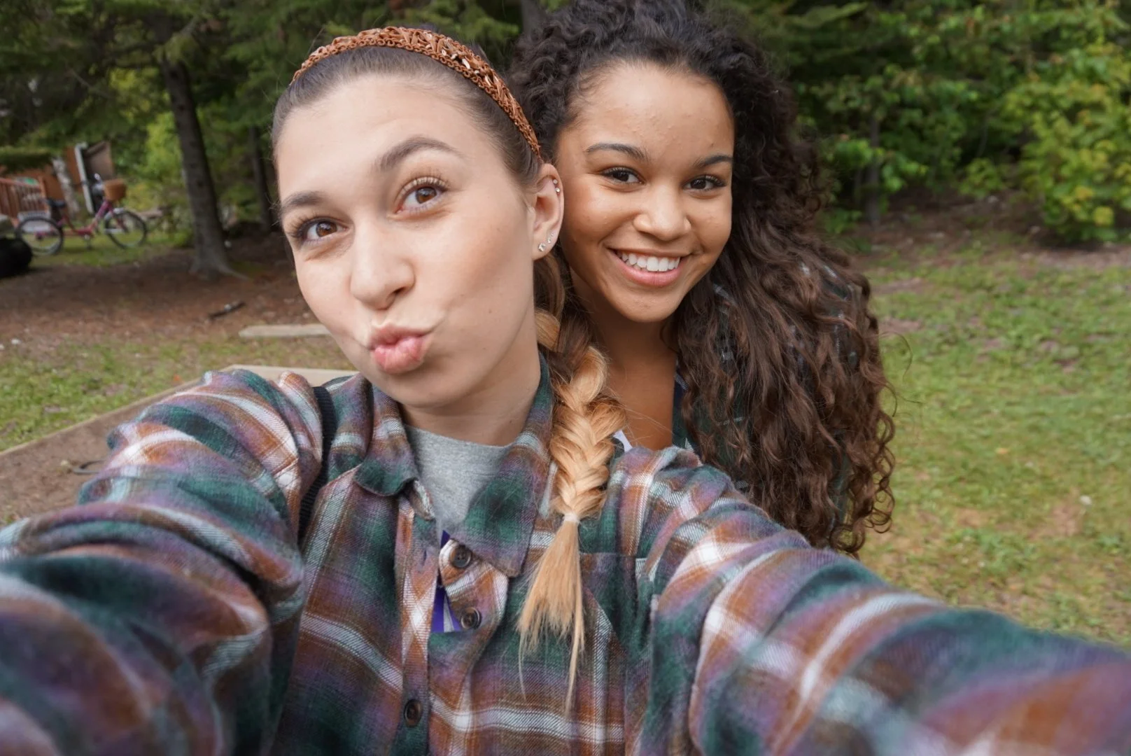 Two young women taking a selfie outdoors in a park with trees in the background. One woman has light skin and brown hair in a braid, making a pouty face, and the other has darker skin and curly hair, smiling.