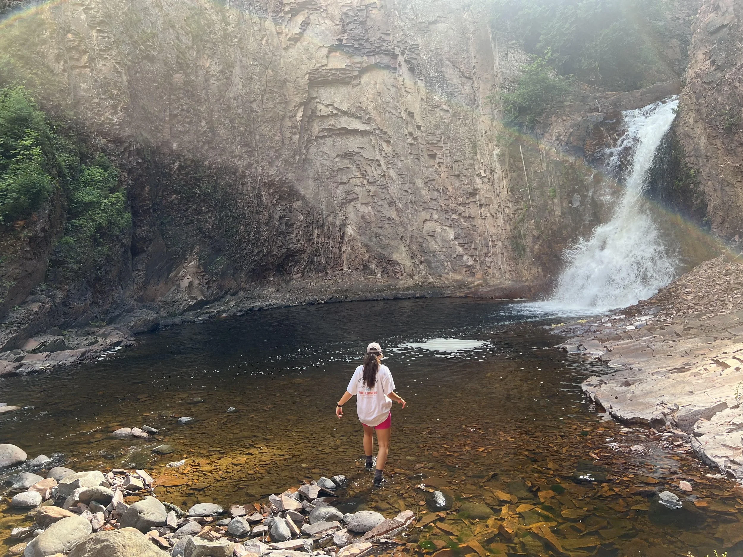 A person wading through a shallow creek near a waterfall surrounded by large rocks and high canyon walls.