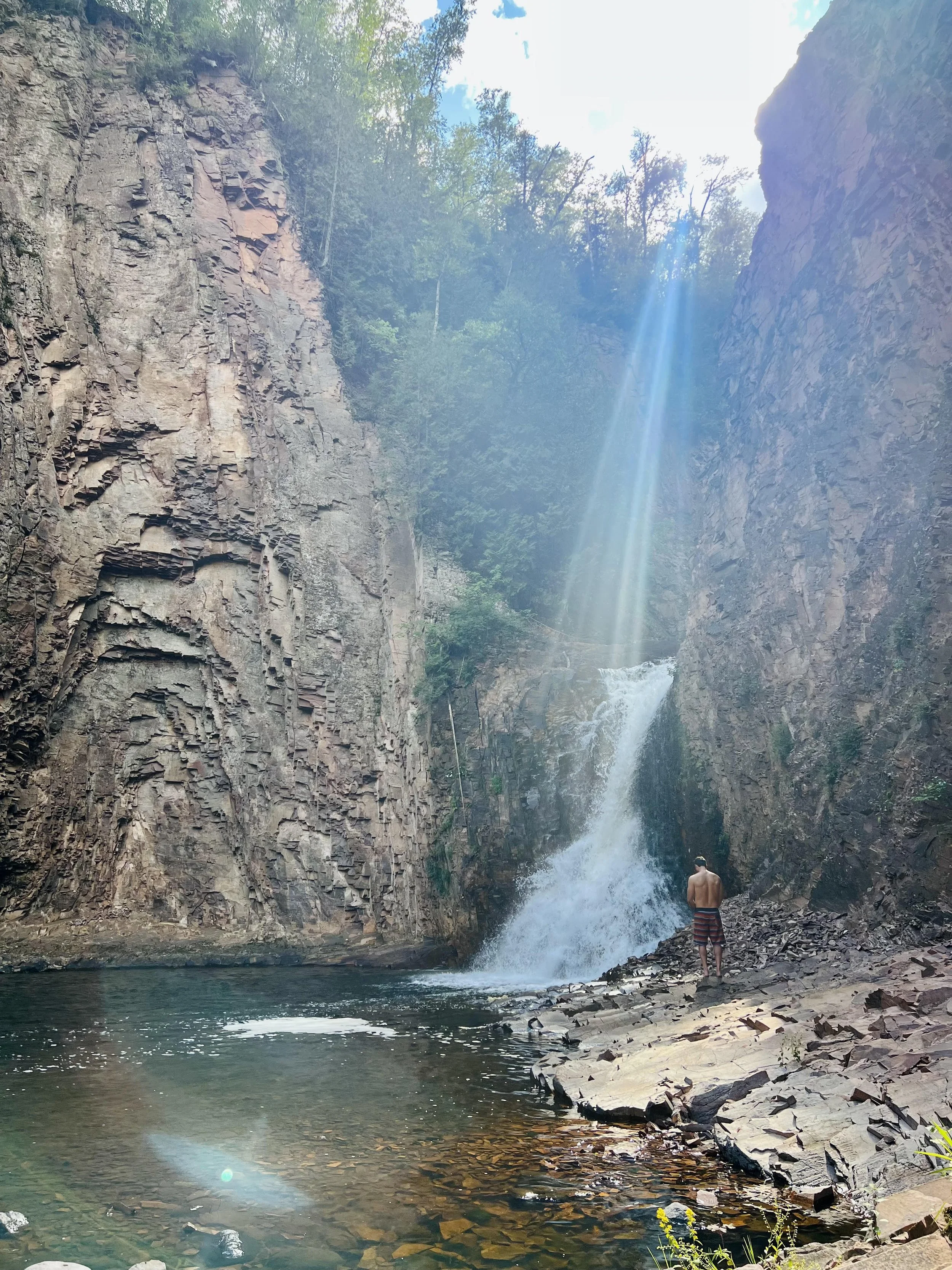 A person stands on rocks beside a waterfall cascading down a rocky cliff into a shallow pool, with a beam of sunlight shining through the trees above.