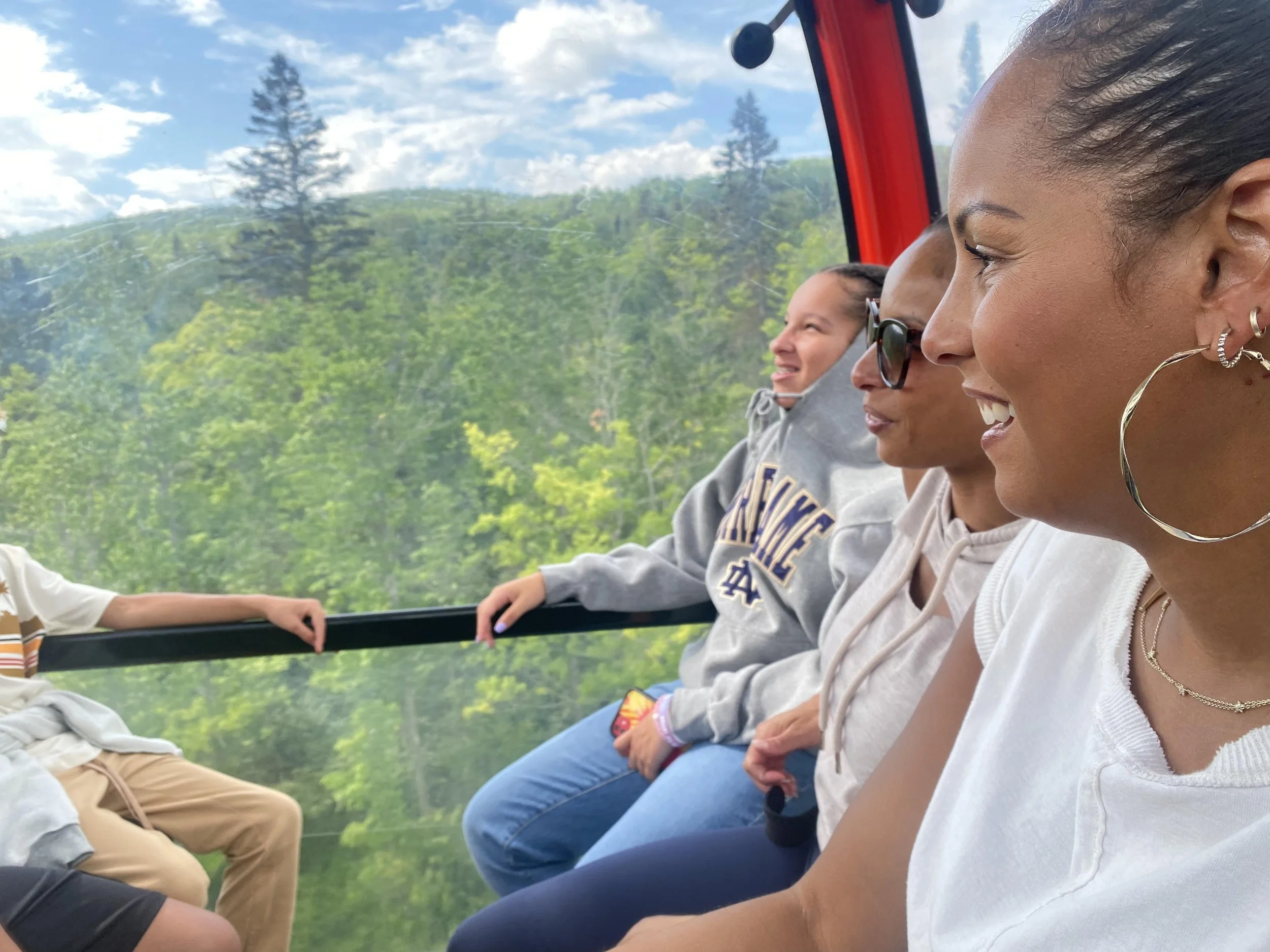 Three women and a child sitting inside a cable car with large windows, enjoying a scenic view of green trees and a blue sky with clouds.
