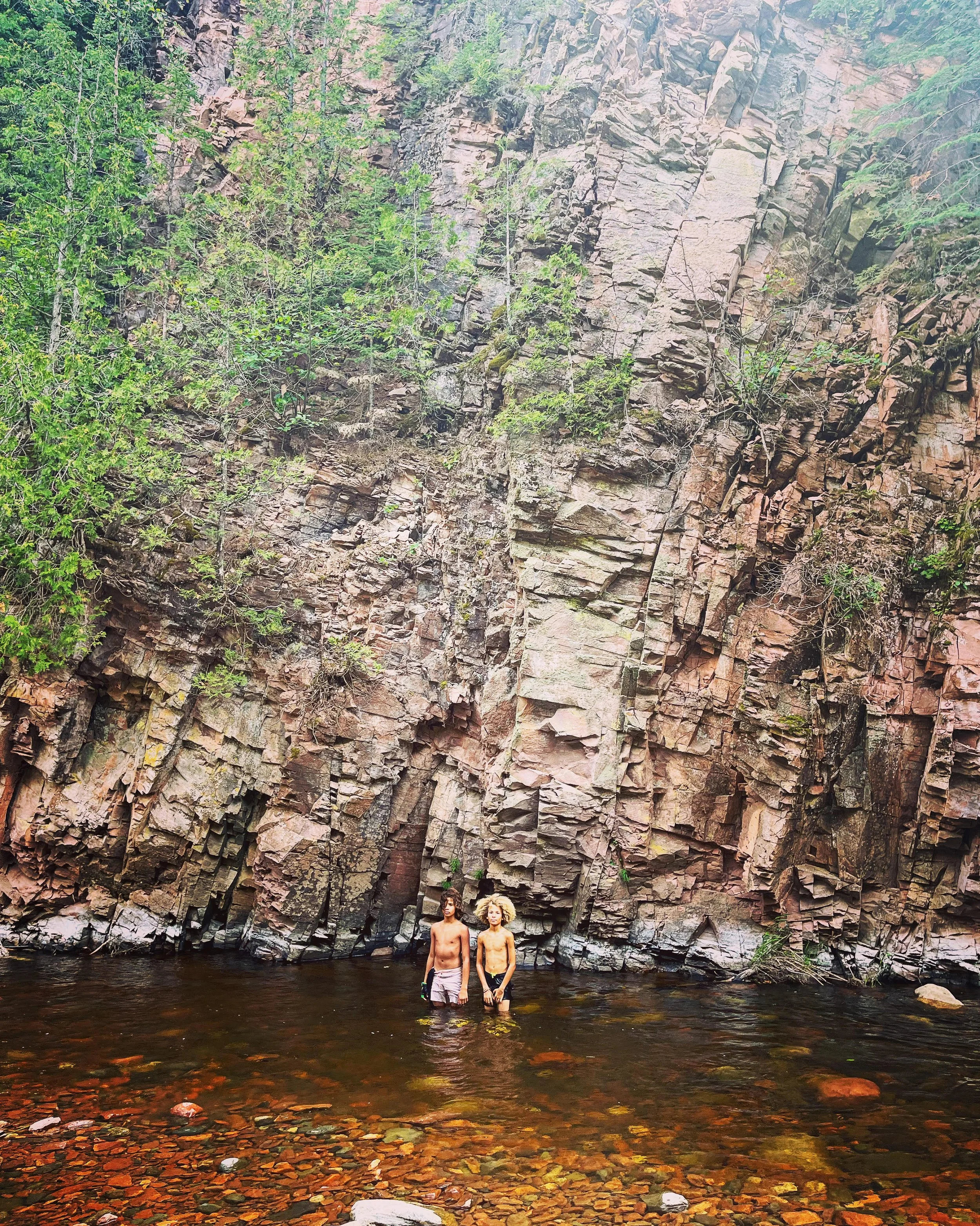 Two shirtless people standing in a shallow river, with a large rocky cliff and trees in the background.