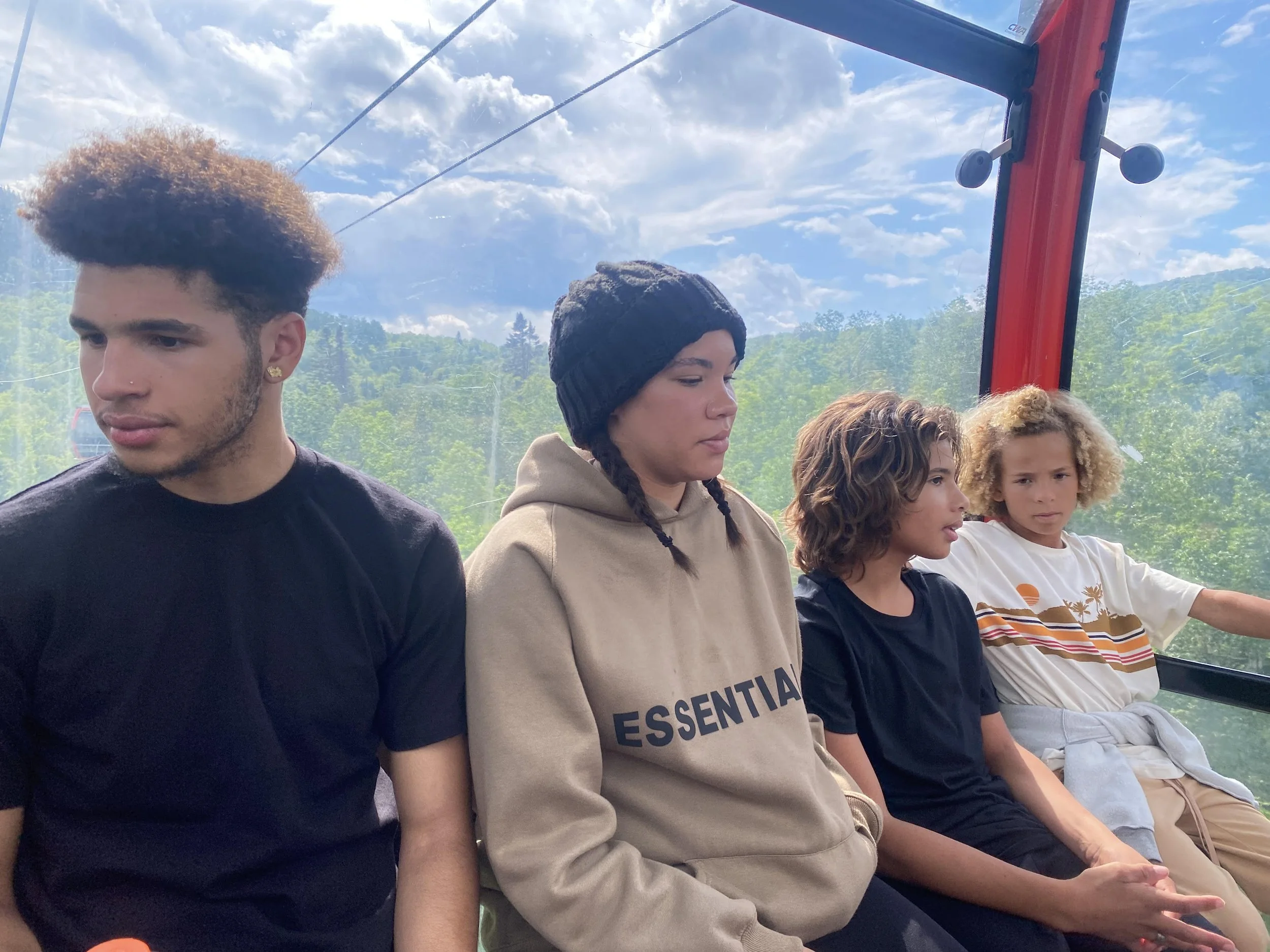 Four diverse young people sitting inside a cable car with a view of green trees and a cloudy sky outside.
