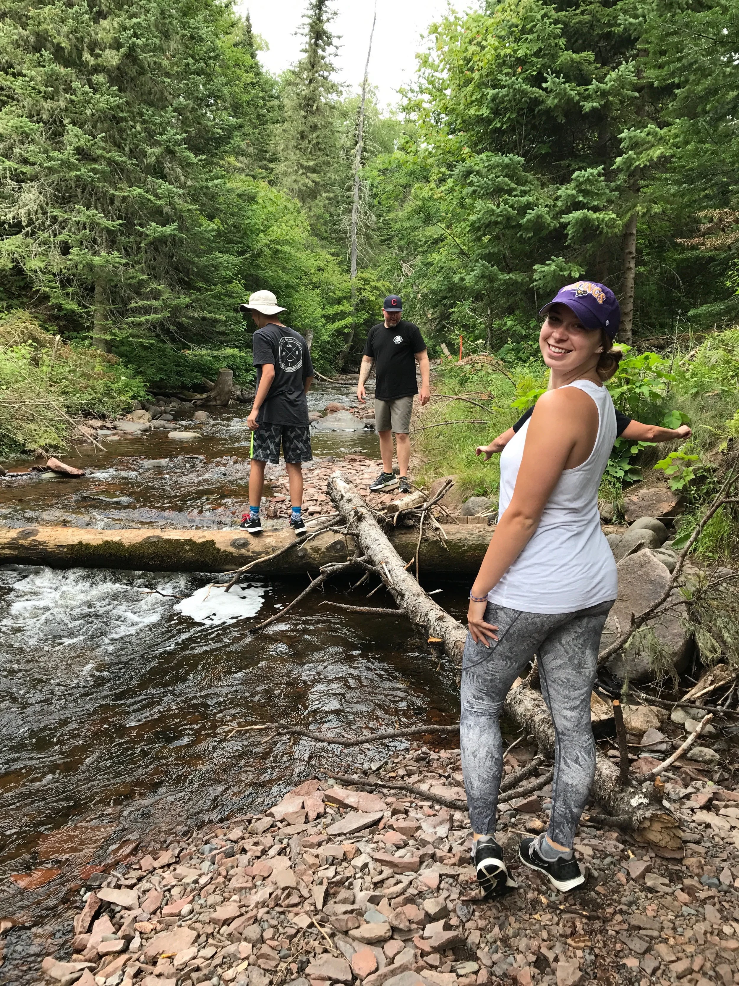 A group of four people in a forest by a creek, with one woman smiling at the camera, three others walking across a fallen log over the water, surrounded by green trees and rocks.