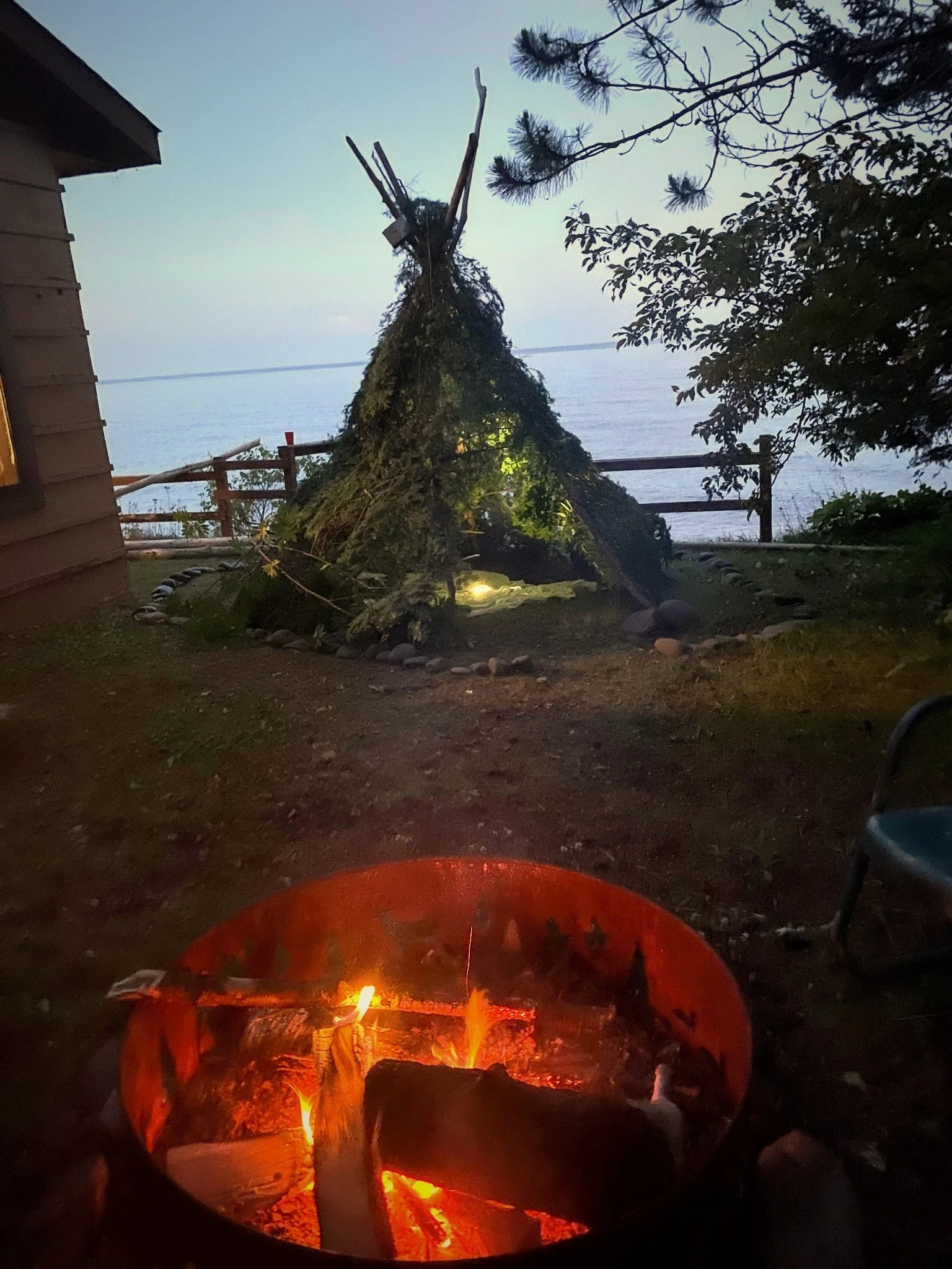 A campfire with burning logs in a fire pit is in the foreground, with a backyard patio leading to a view of a lake in the background. Nearby are trees and a wooden fence, and a triangular teepee-like structure made of branches and foliage is position
