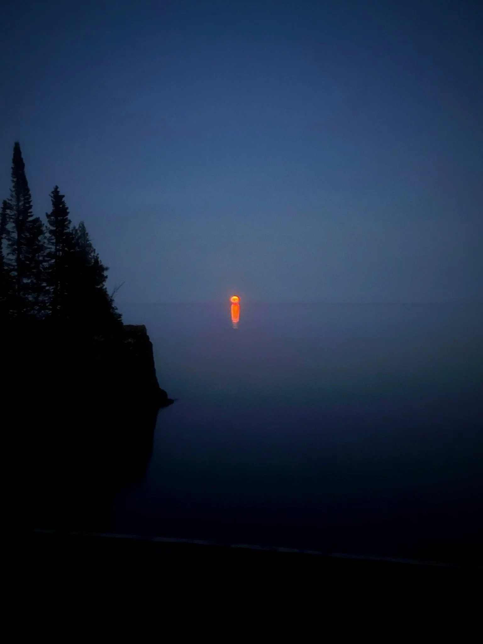 A gradient sunset or sunrise over a calm body of water with a silhouetted tree-lined shoreline on the left and a glowing orange sun near the horizon reflected in the water.