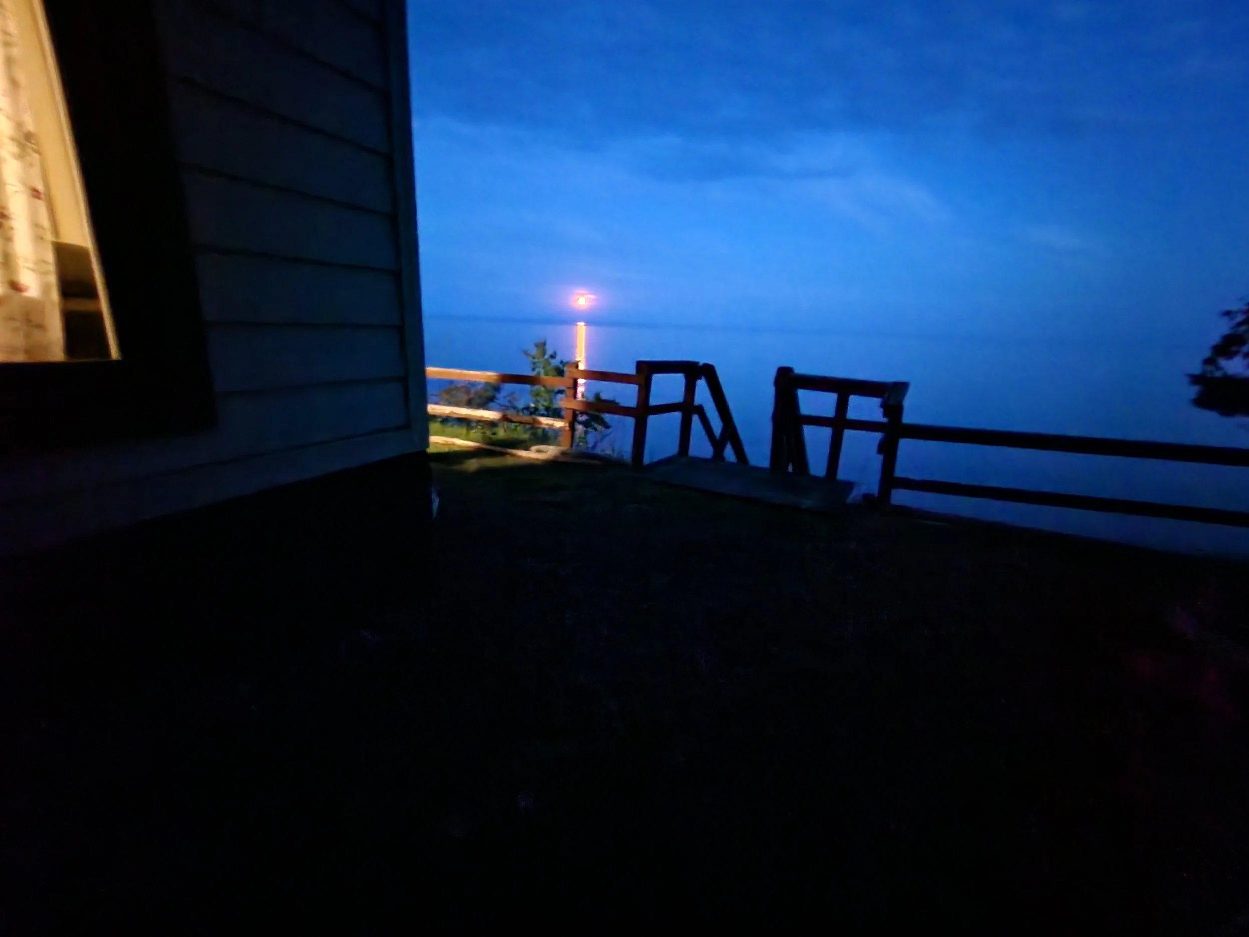 A dimly lit outdoor wooden deck or balcony at night, with a view of the ocean and a bright moon or lighthouse on the horizon.