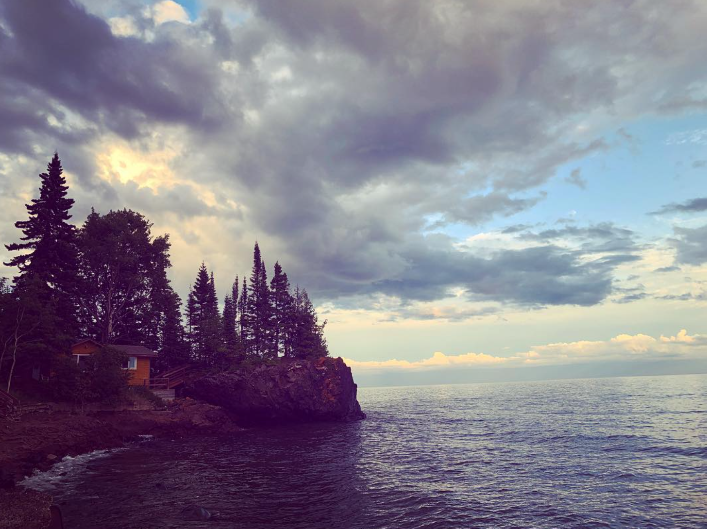 A coastal scene with a rocky shoreline, evergreen trees, and a house on the left, under a partly cloudy sky.