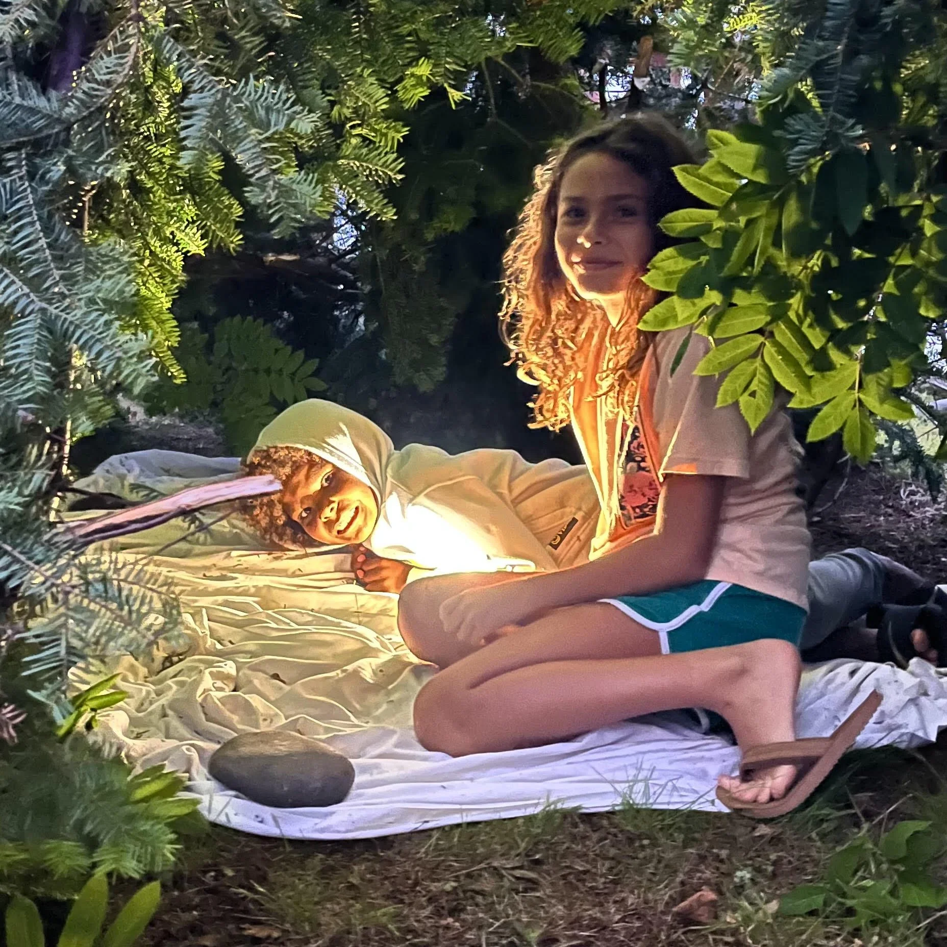 Two children, a boy and a girl, are inside a small makeshift tent made of fabric and greenery, illuminated by a light source. The boy is lying on his side, smiling and wearing a hoodie, while the girl is sitting on the ground, smiling and looking at 