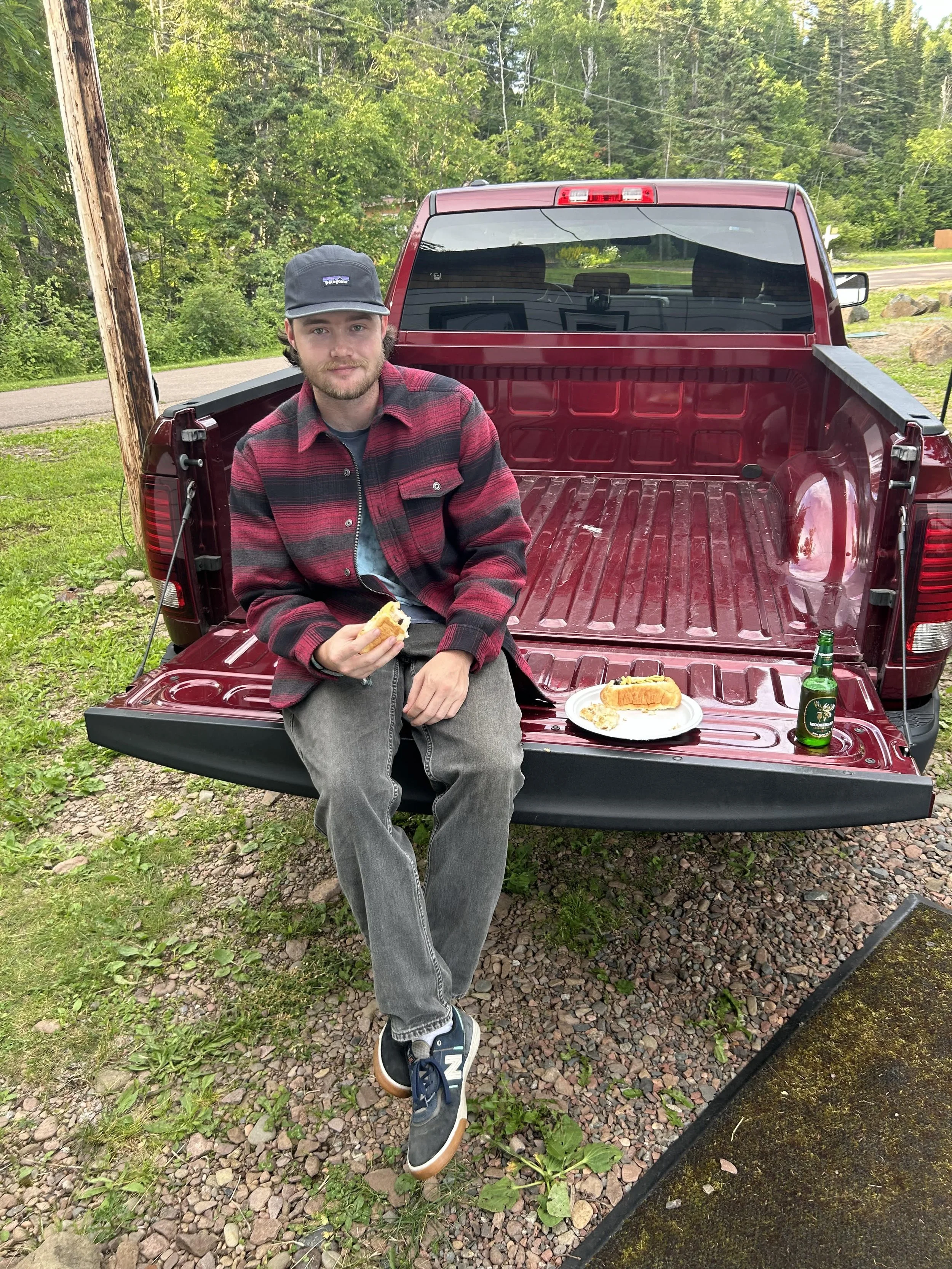 Young man sitting on the open tailgate of a red pickup truck, eating a hot dog. There is a plate with food and a beer bottle on the tailgate. Green trees and a rural road in the background.