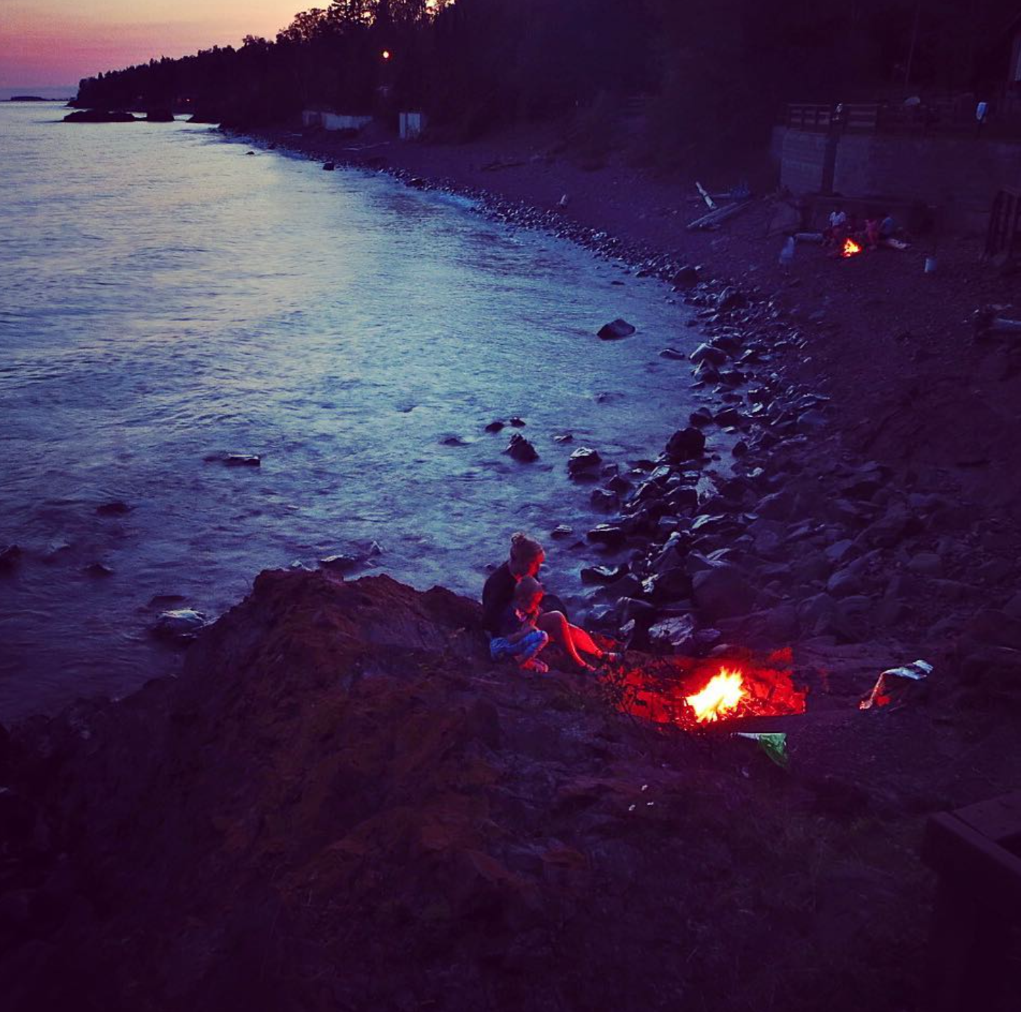 Two children sitting on rocks by a lakeshore at sunset, with a small campfire burning nearby, and trees in the background.