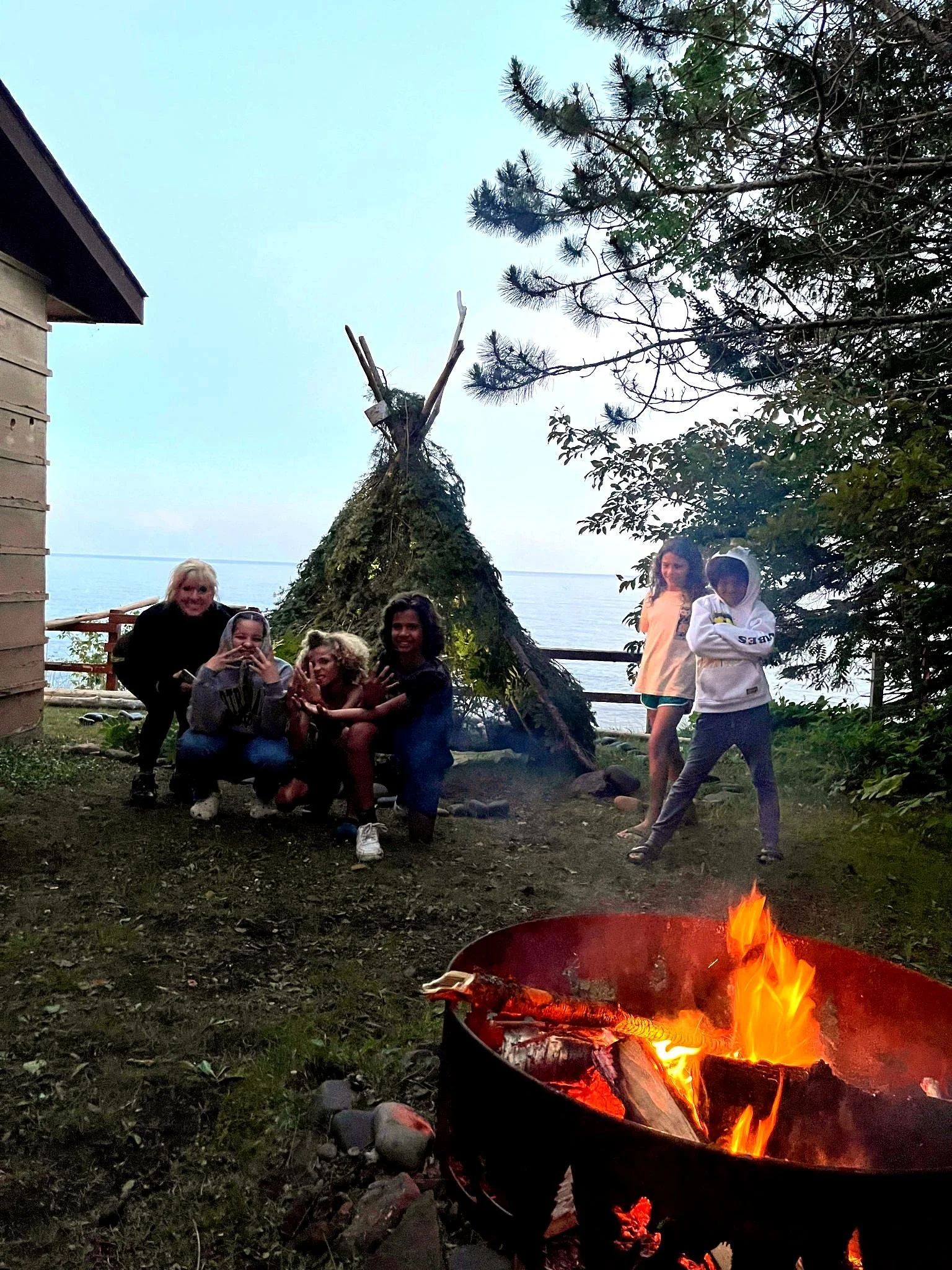 Children gathered around a campfire on a grassy area near a wooden teepee structure and trees, overlooking a body of water at dusk.