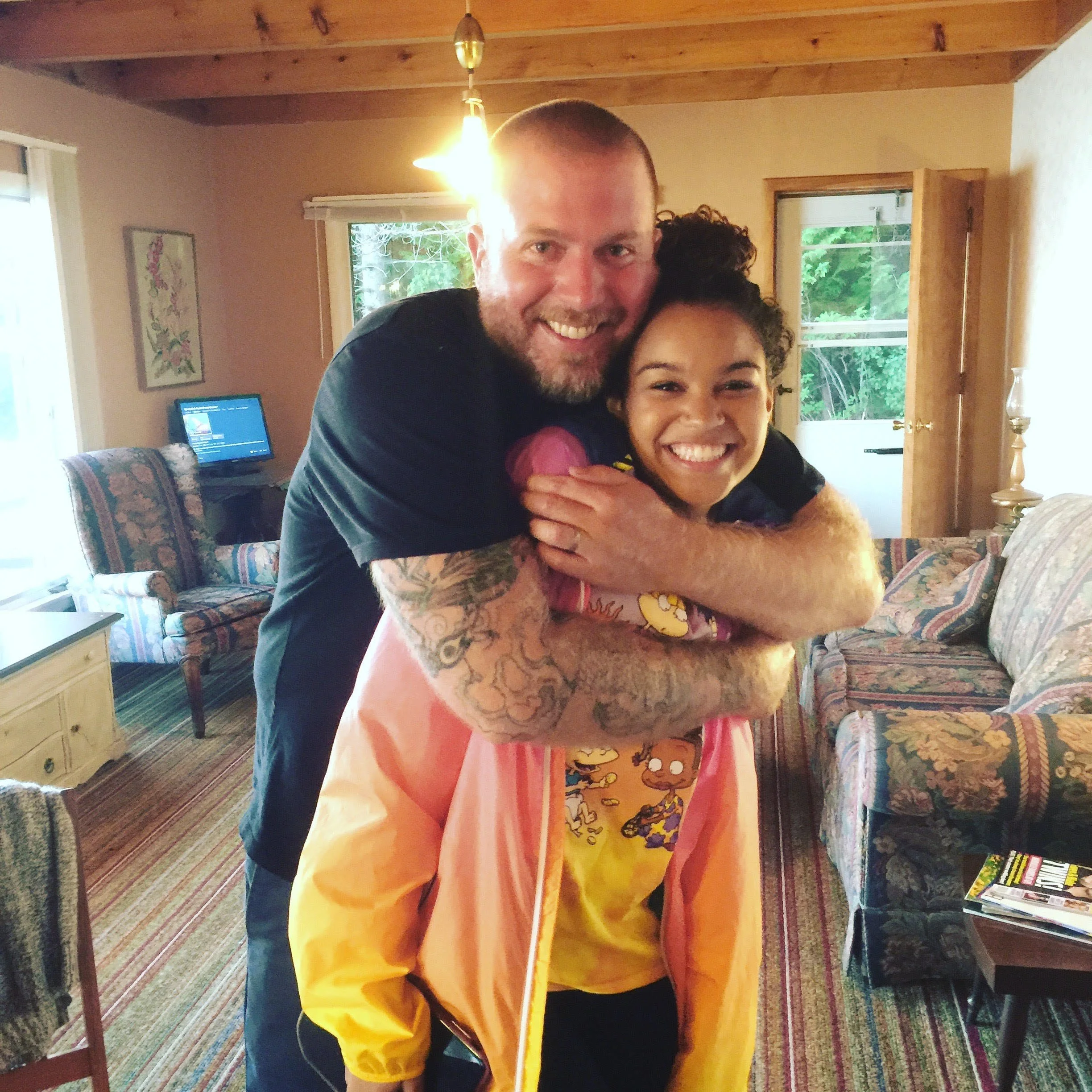 A man with tattoos hugging a smiling girl in a colorful room with patterned furniture.