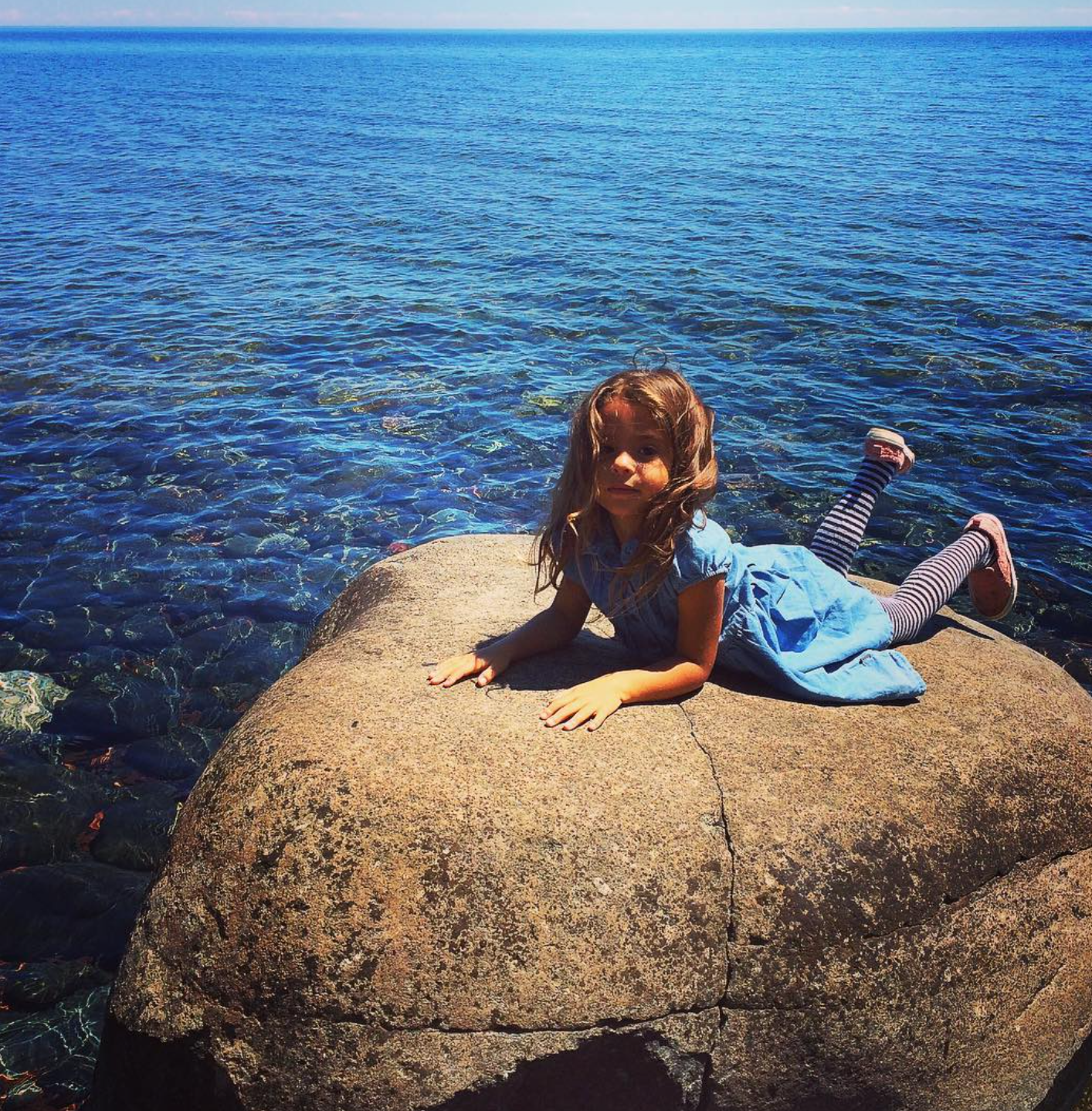 A young girl with long brown hair is lying on her stomach on a large rock by the water, wearing a blue dress and striped leggings with sandals, near a body of water on a sunny day.