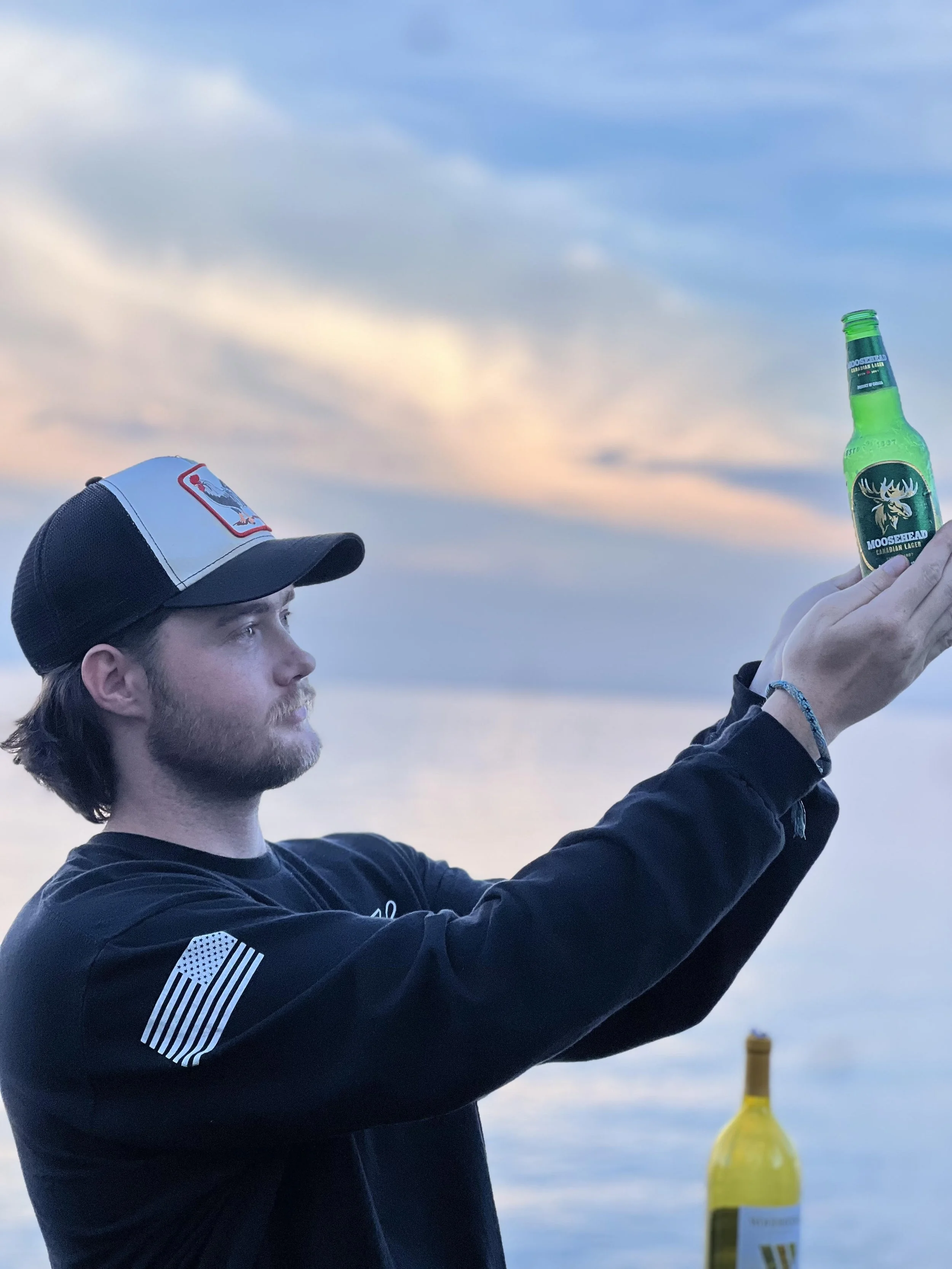 A man with a beard, wearing a black sweatshirt with a flag patch on the sleeve and a trucker hat, is holding a green bottle of Moosehead beer with both hands against a sunset sky.