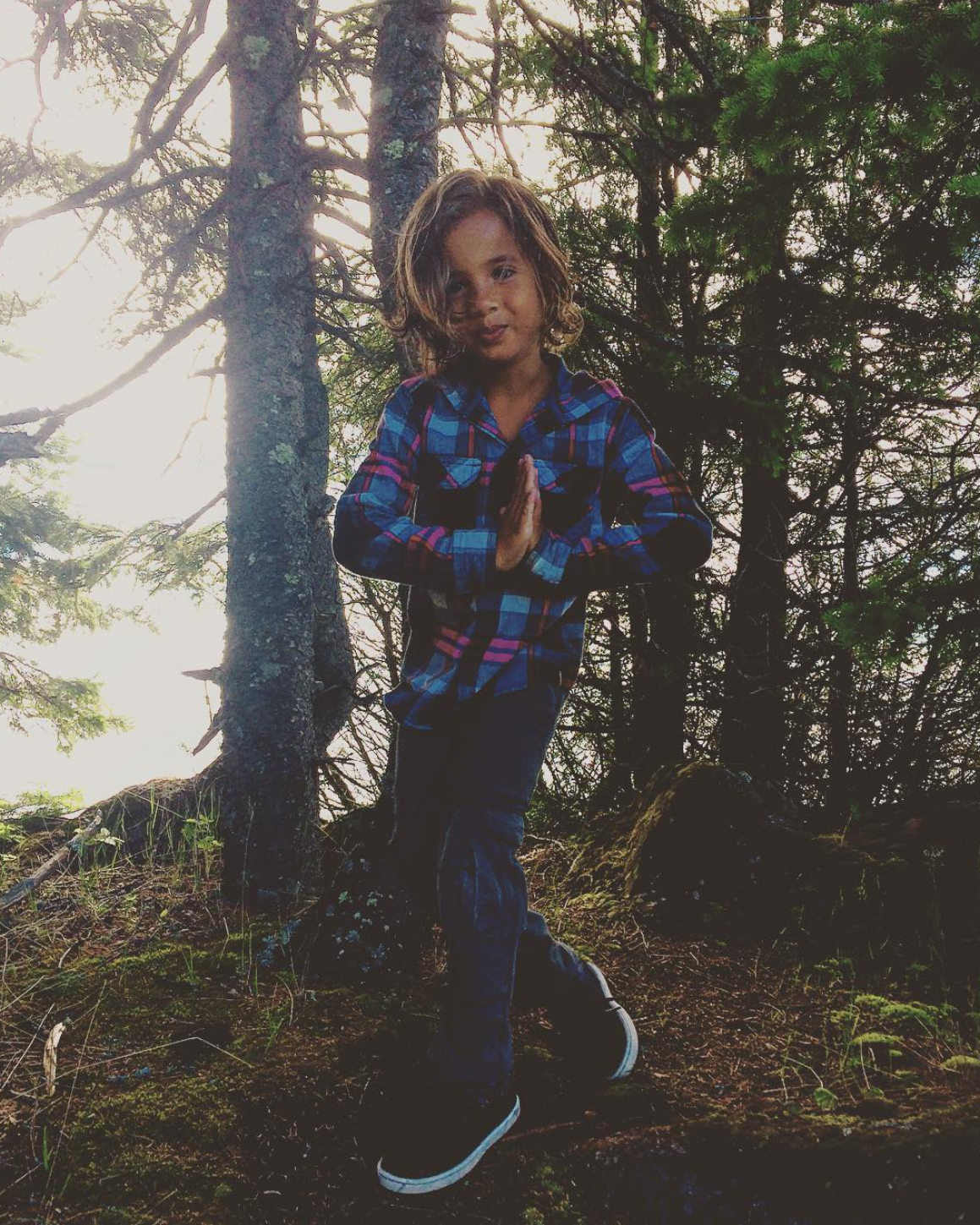 Child in a plaid shirt standing on a forest trail, surrounded by trees, with hands together in a prayer or greeting gesture.