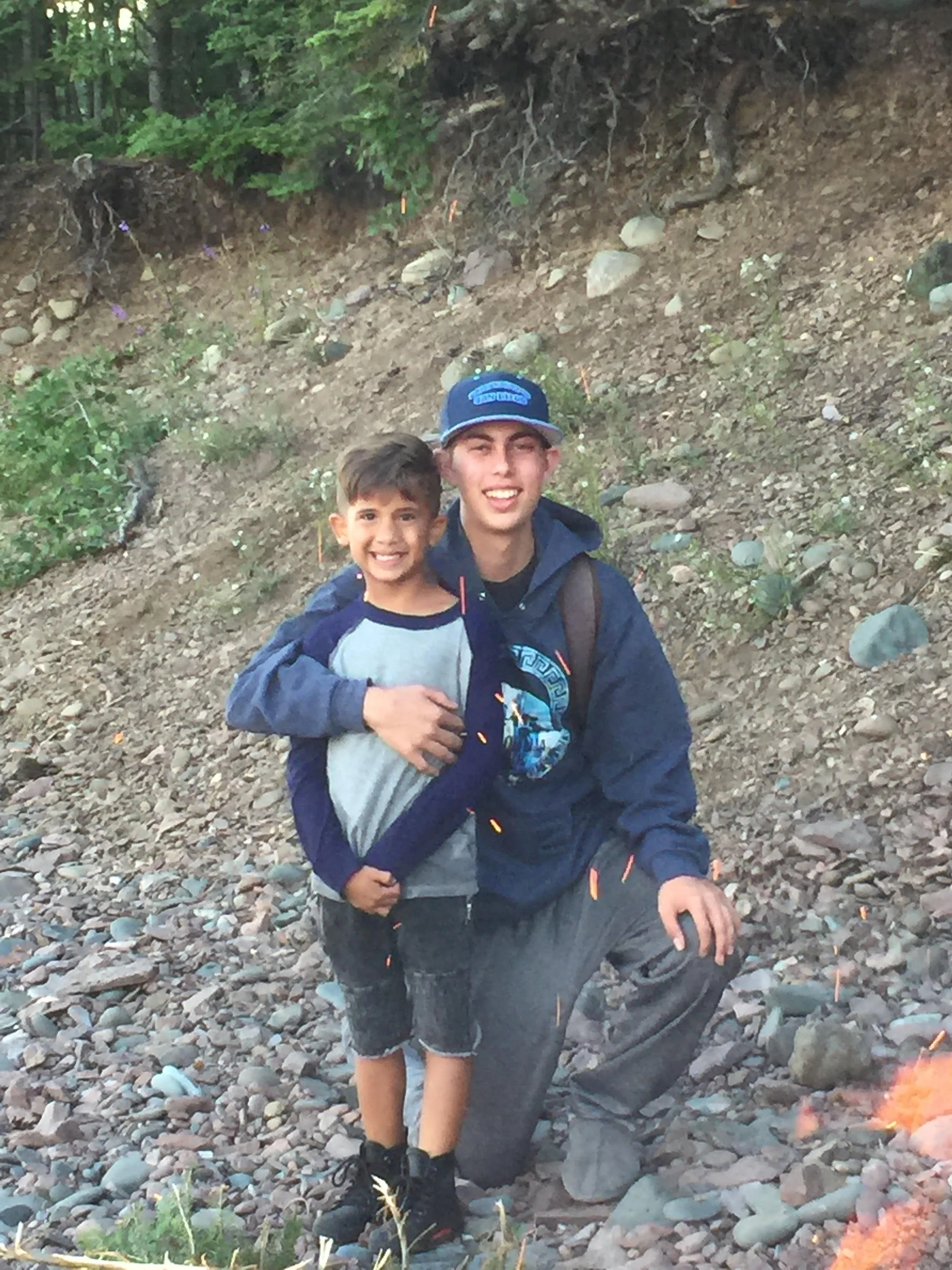 A young boy and a teenage boy are smiling and posing together outdoors on a rocky slope surrounded by trees.
