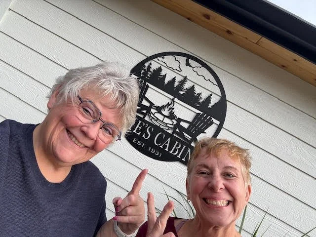 Two smiling women with short gray hair standing outside next to a white house with horizontal siding, in front of a black metal sign that reads "Louise's Cabin" with trees and a campfire illustration.