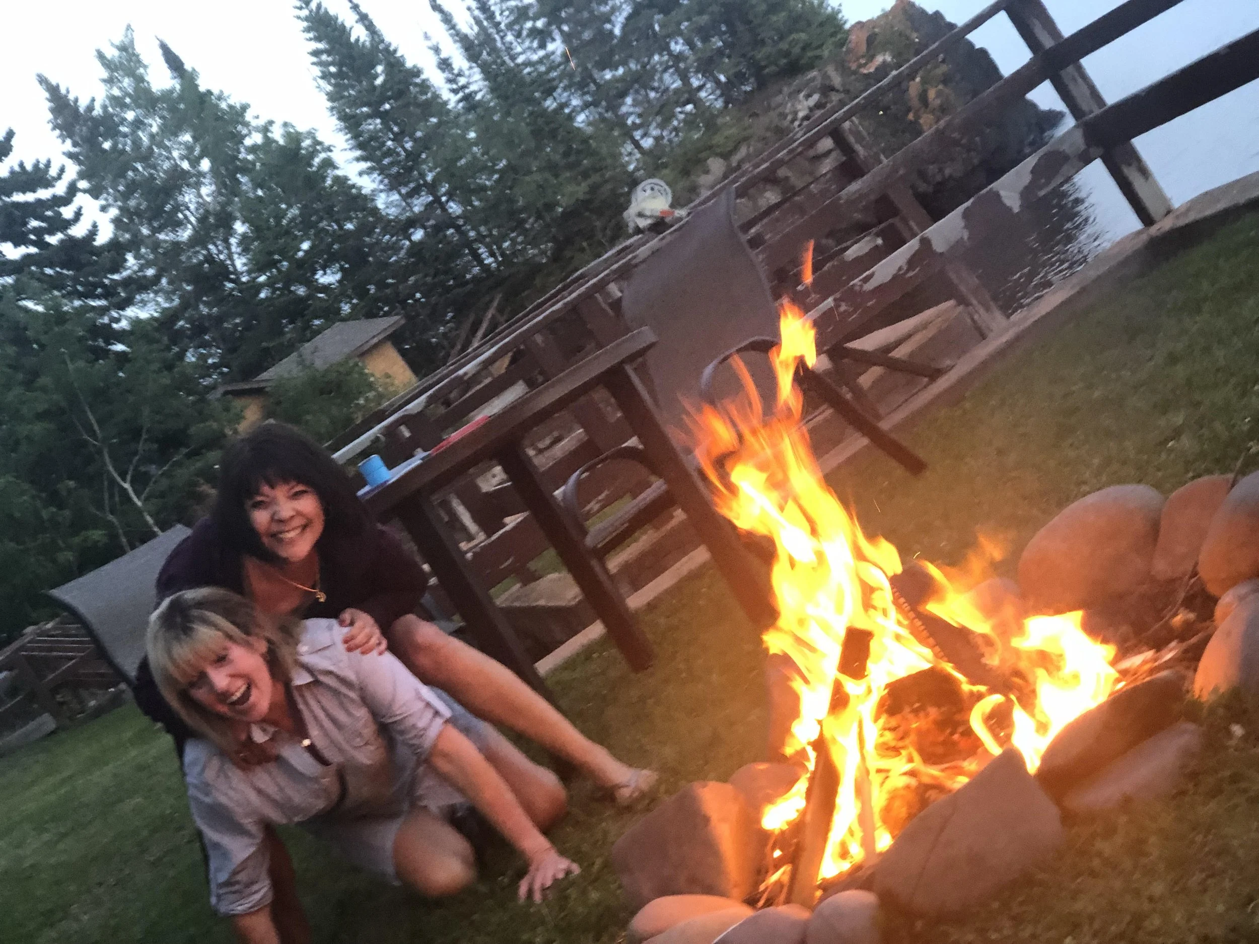 Two women happily crouching beside a campfire outdoors at twilight with trees, a fence, and a building in the background.
