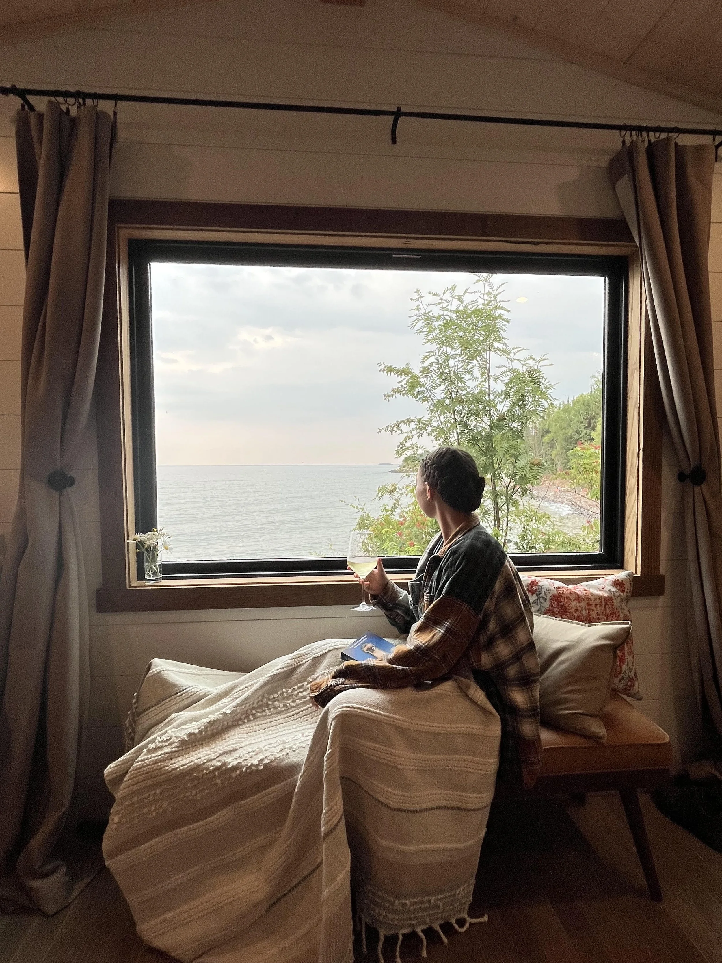 Person sitting on a cozy bed with a blanket, holding a glass of white wine, looking out the window at a body of water and green trees outside.