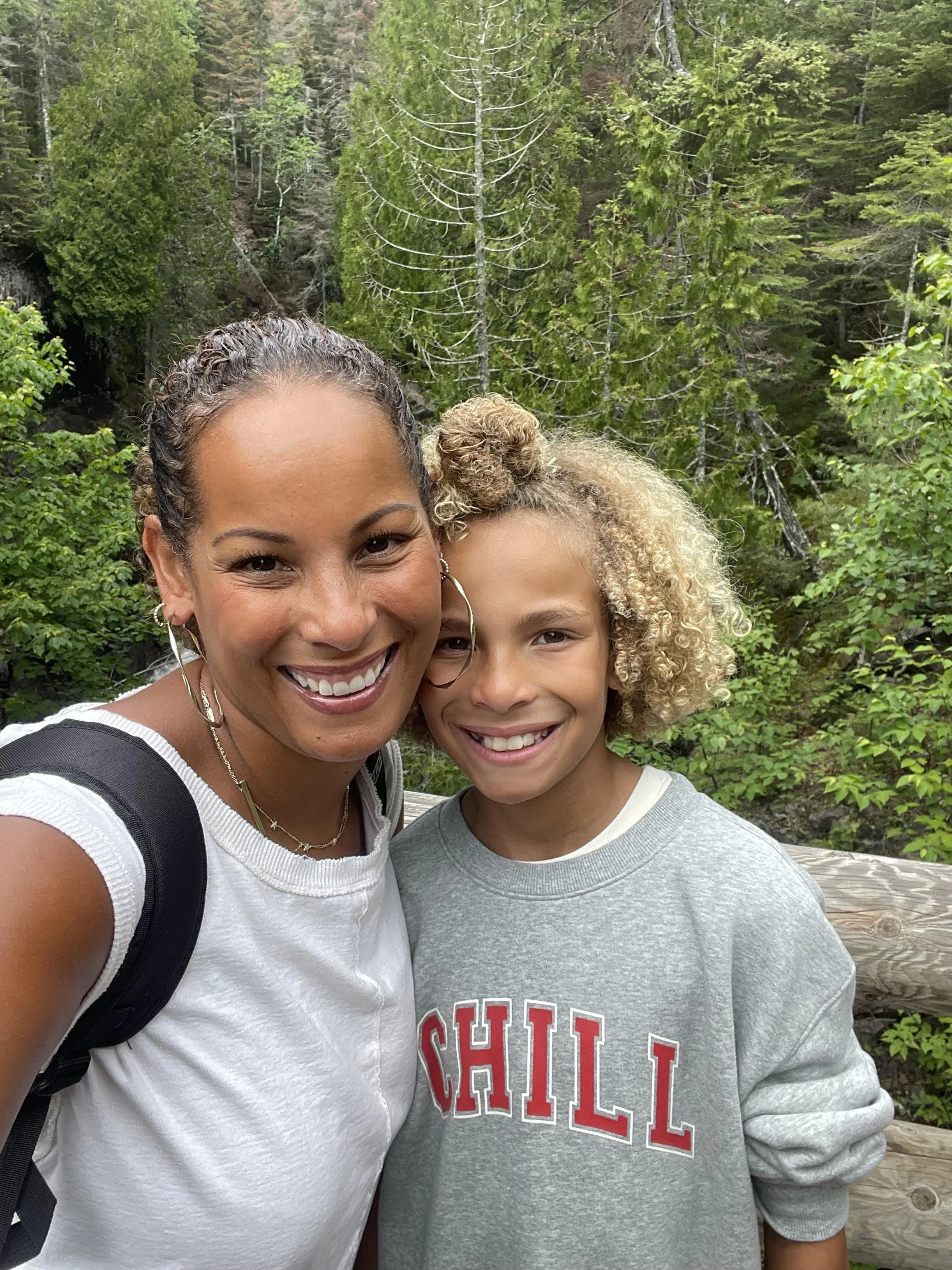 A woman and a young girl smiling and taking a selfie in a forested area with green trees and wooden fence in the background.