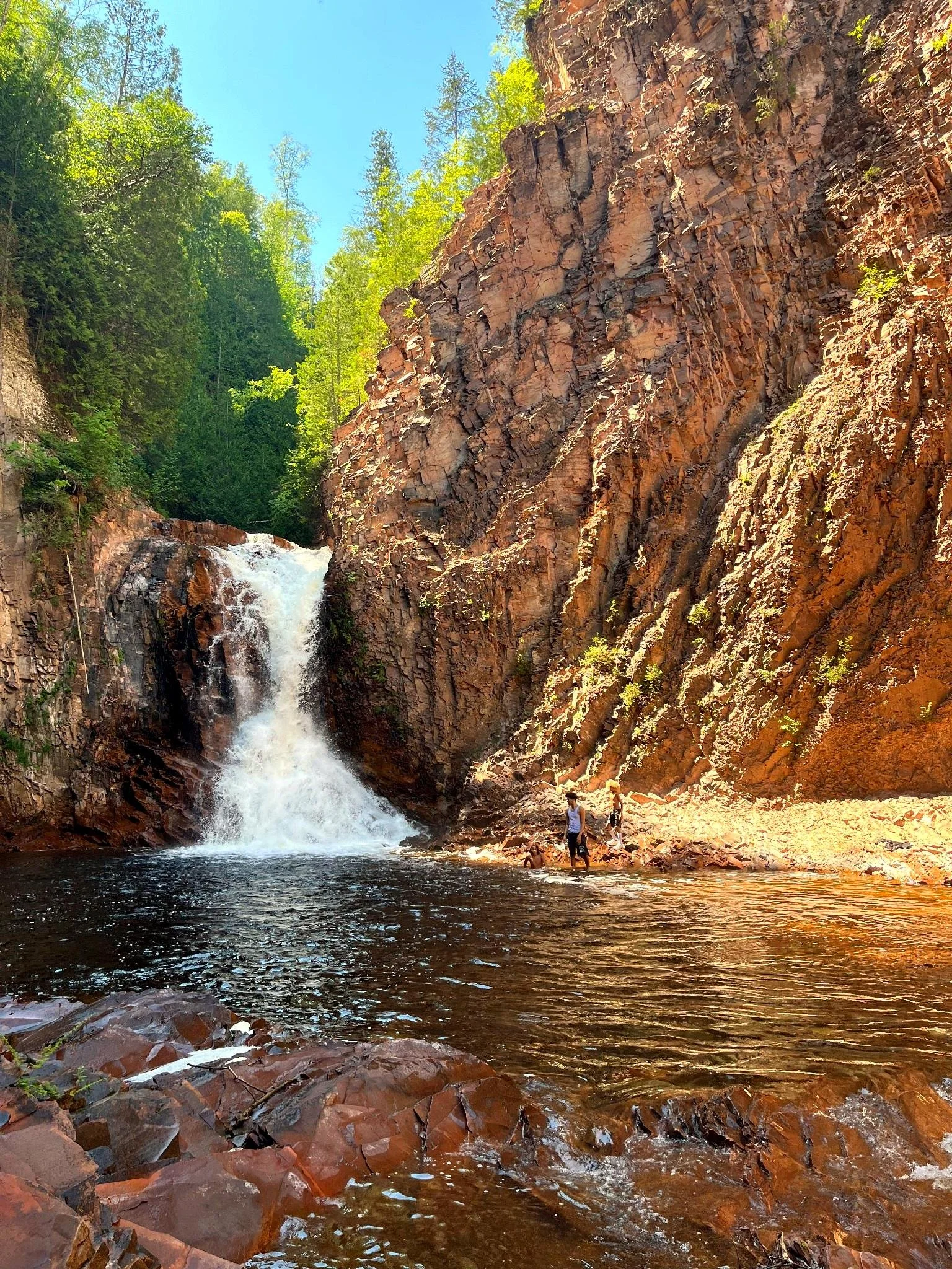 A scenic view of a waterfall cascading into a pool surrounded by rocky cliffs and lush green trees, with a clear blue sky overhead.