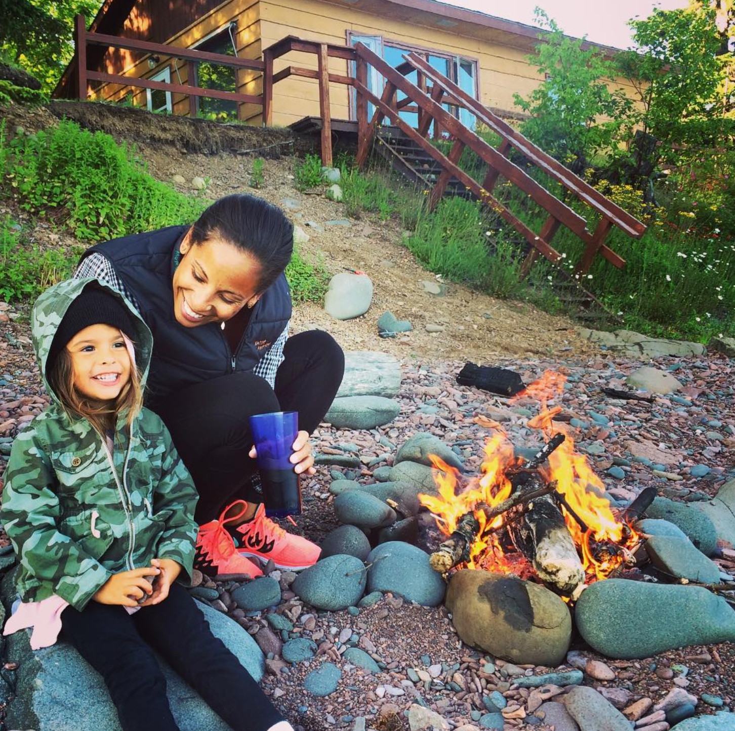 A woman and a girl sitting next to a campfire on a rocky beach, smiling and enjoying the outdoor scene, with a house and greenery in the background.