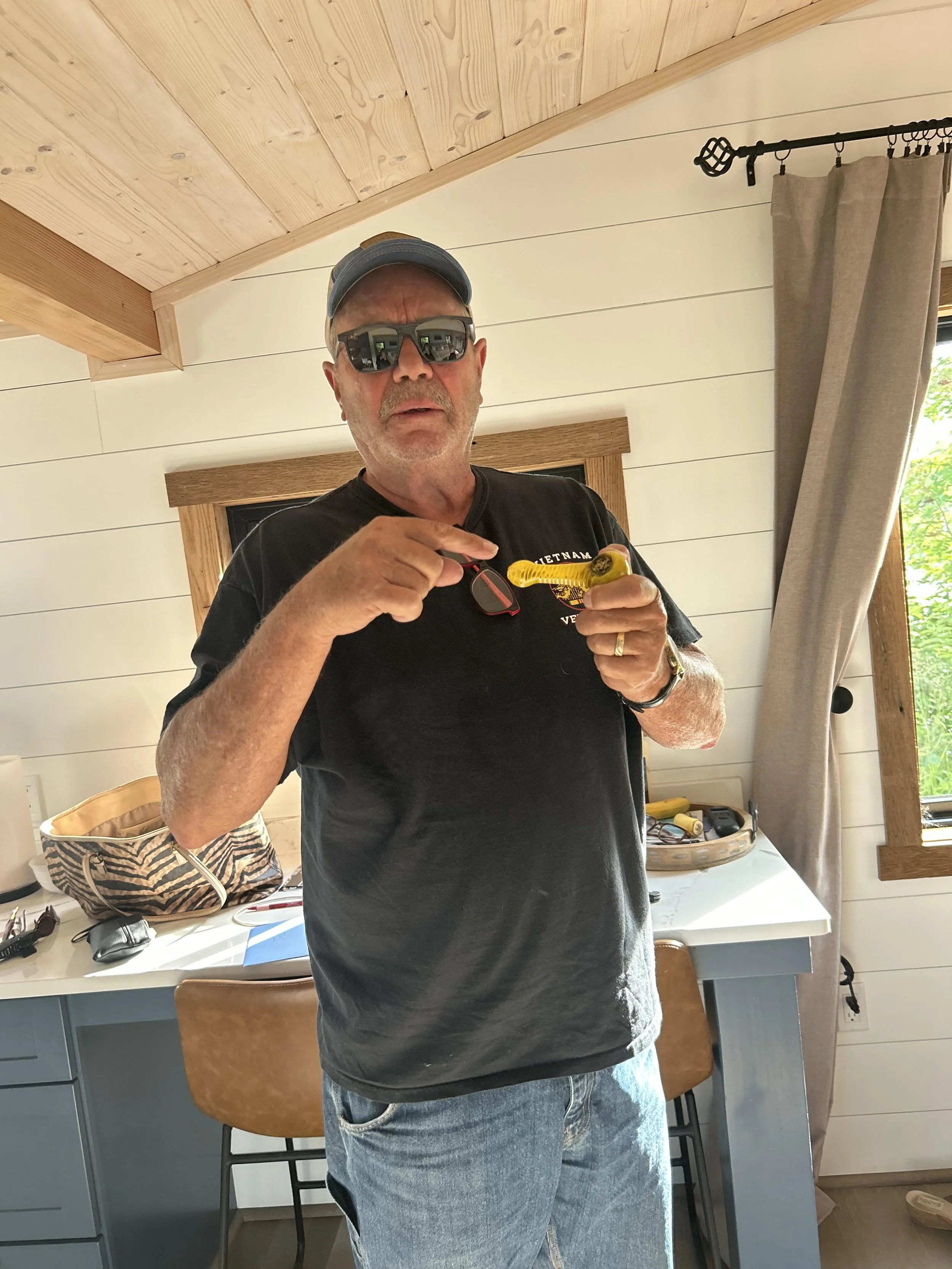 A man wearing a black t-shirt, blue jeans, sunglasses, and a baseball cap stands inside a room with wooden walls and ceiling, holding a yellow tool or device and pointing at it with one finger. There is a white countertop with various items behind hi