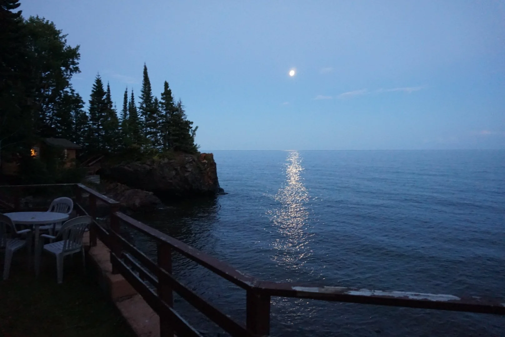 A nighttime view of a lake with the moon reflecting on the water. To the left, there are tall trees and a small house or cabin with a lit window. In the foreground, there is a deck with a few plastic chairs and a small table.