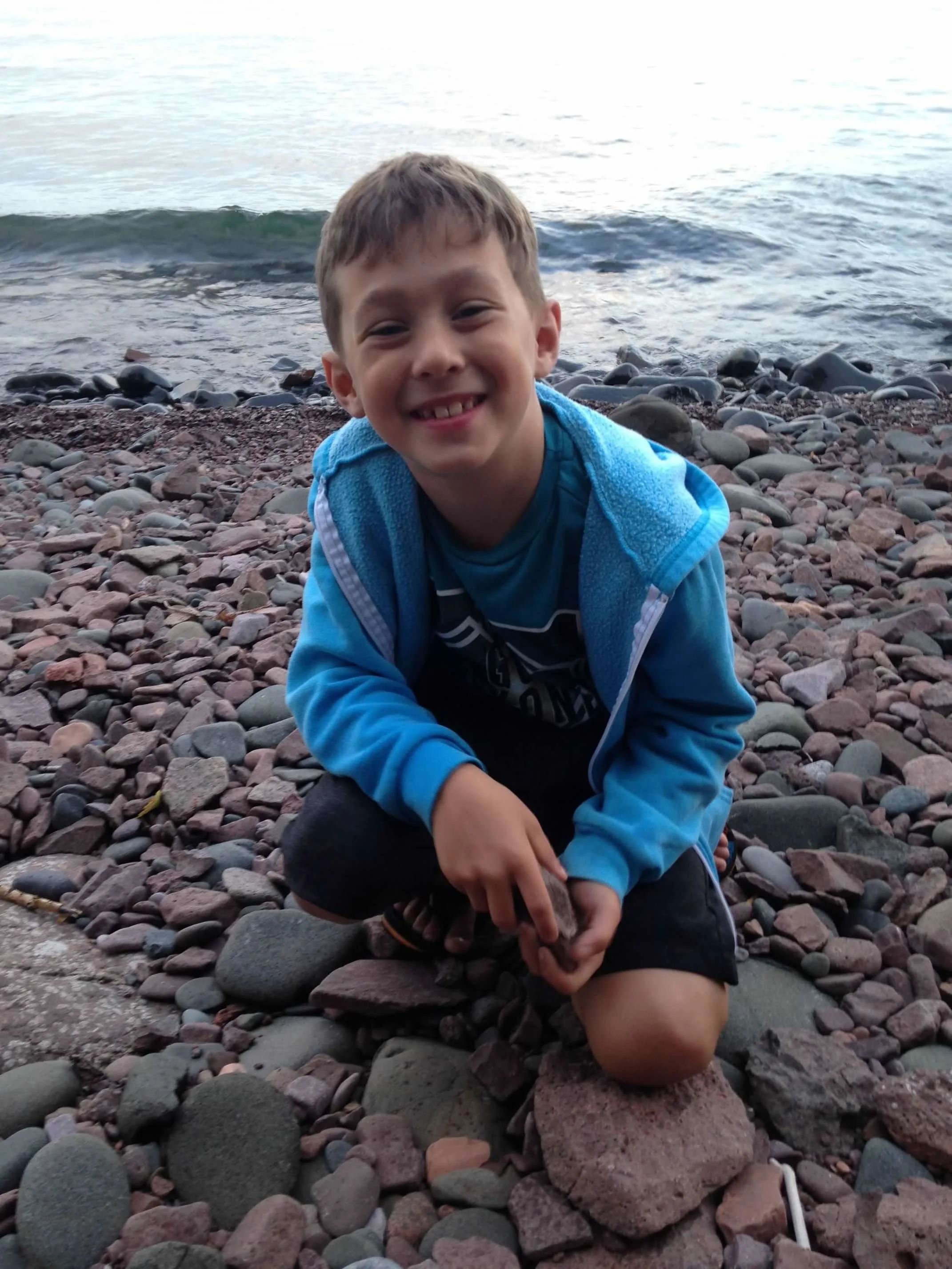 A young boy crouching on a rocky beach near the water, smiling at the camera.