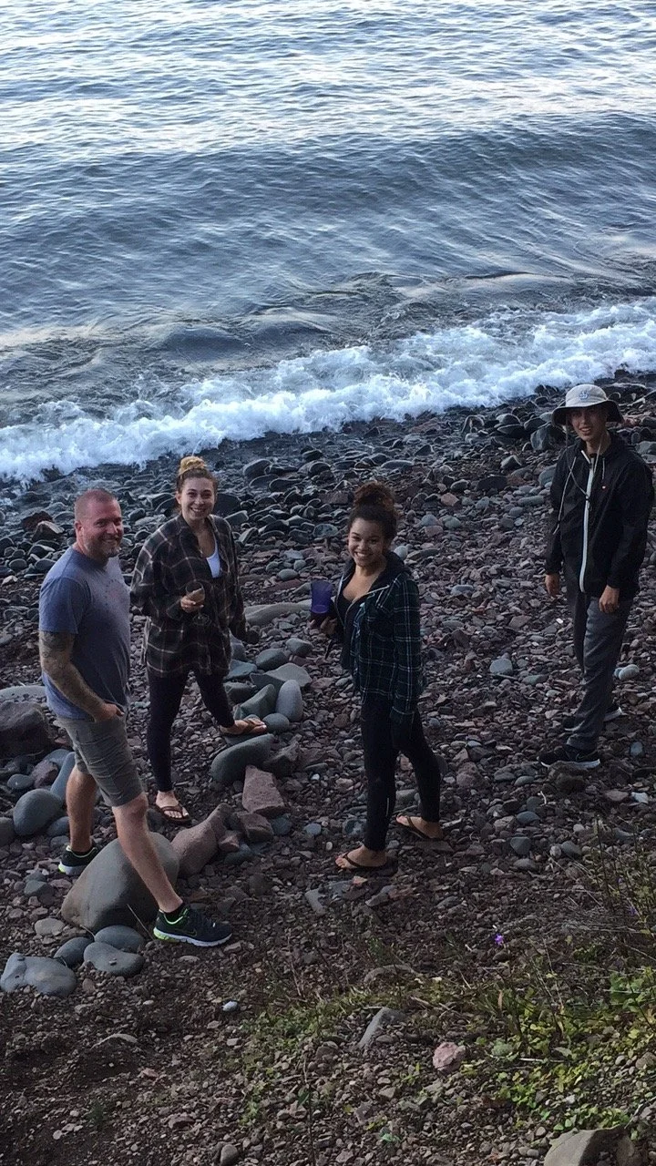 Four people standing on a rocky beach near the ocean waves, smiling at the camera, with some holding drinks.
